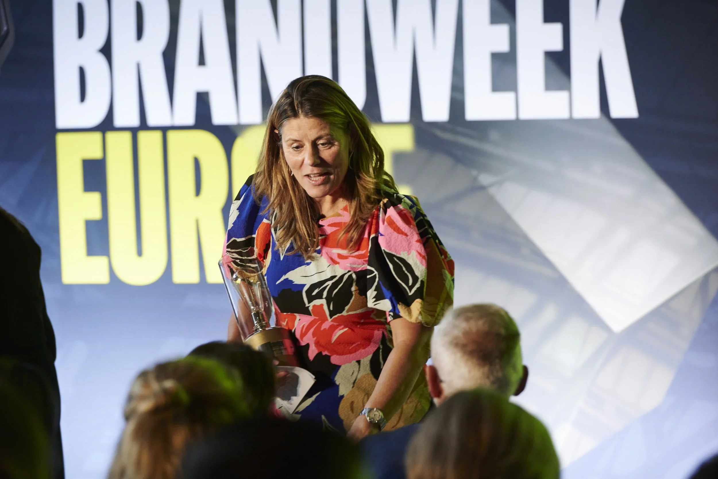 A woman with long brown hair wearing a colorful floral dress holds a trophy, standing in front of a large screen with the words 'BRANTWEEK EUROPE' displayed. She is engaging with a seated audience.
