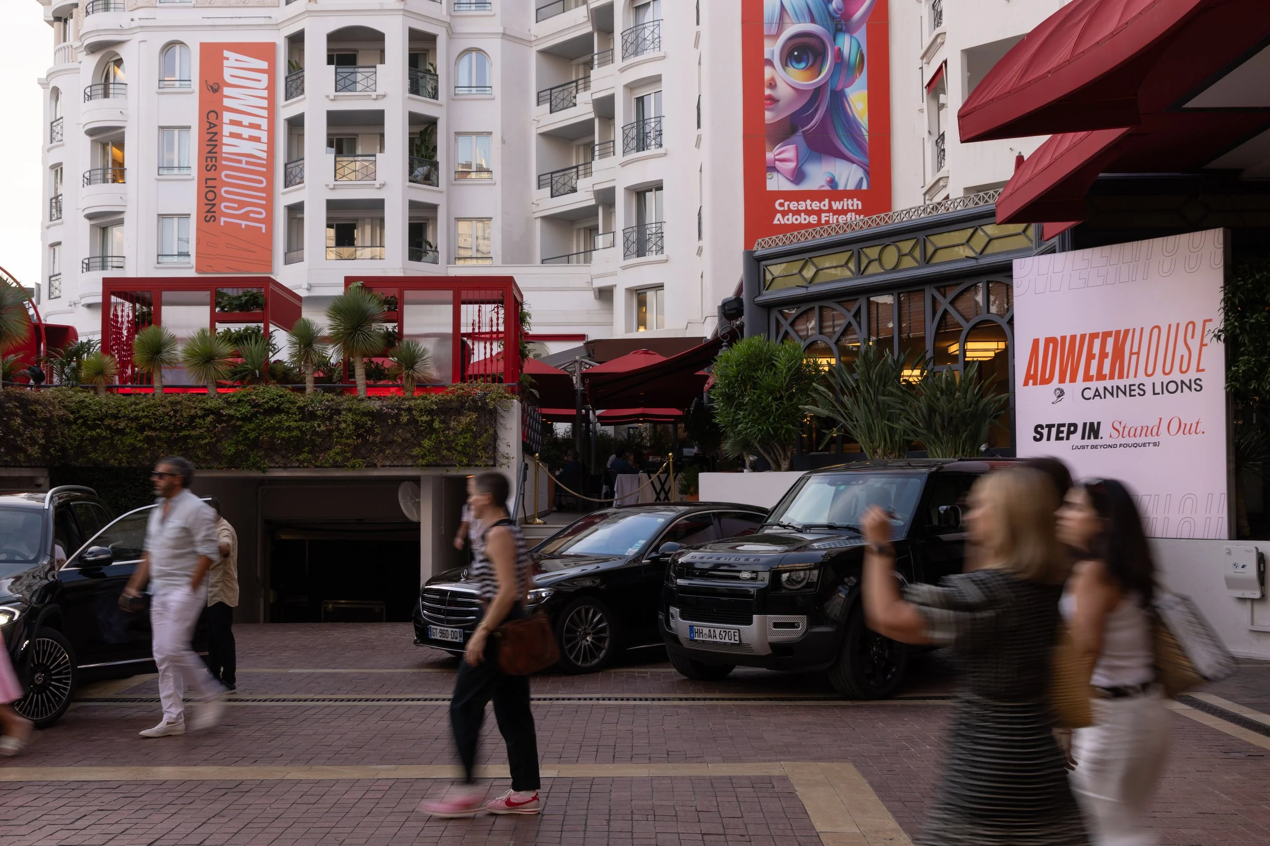 Street scene outside a building with white walls and balconies, featuring advertisements for ADWEEK House Cannes Lions, with several people walking, parked cars, and plants.