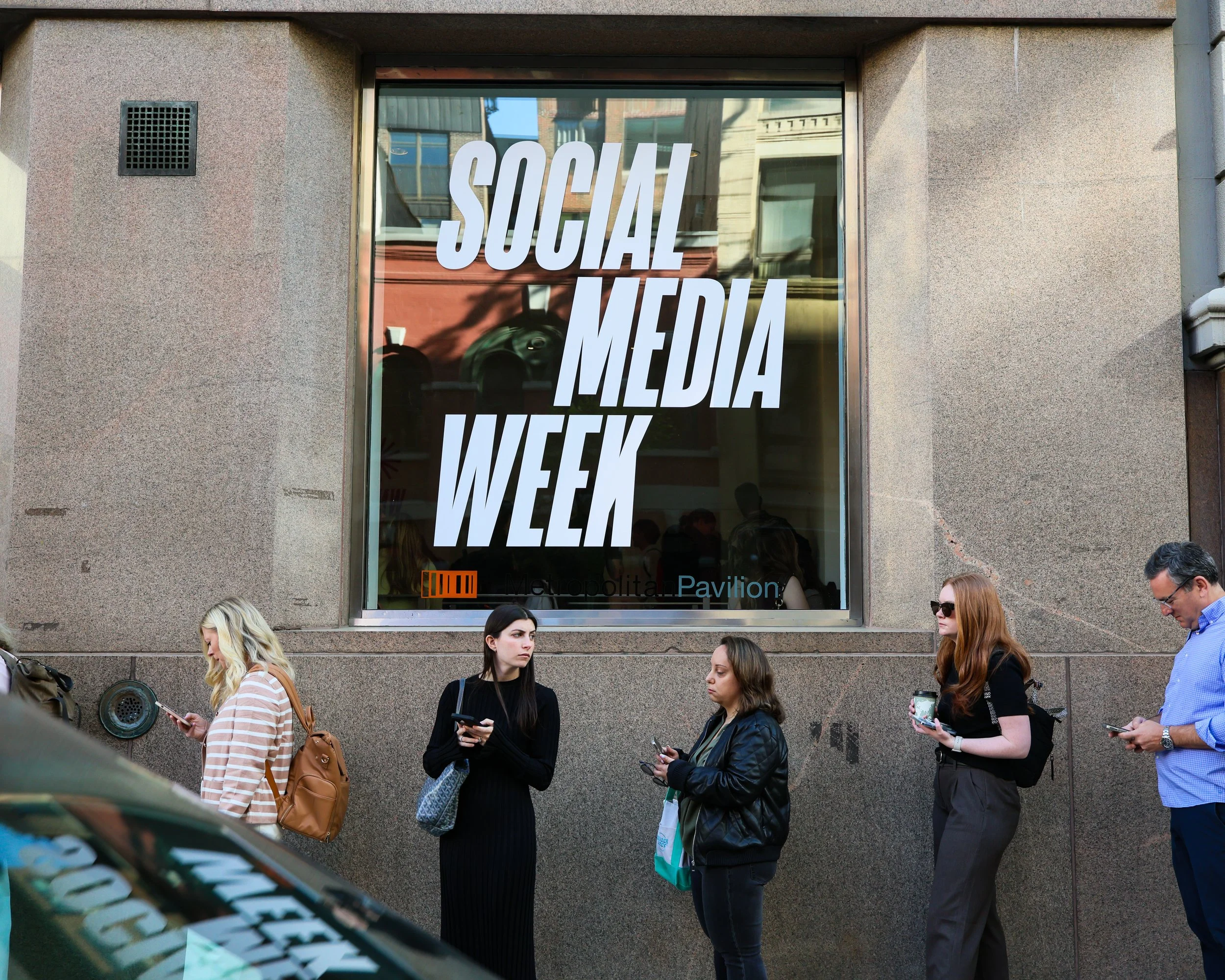 People waiting outside a building during Social Media Week, a large window with bold white text on it, and a reflection of street scene in the window.