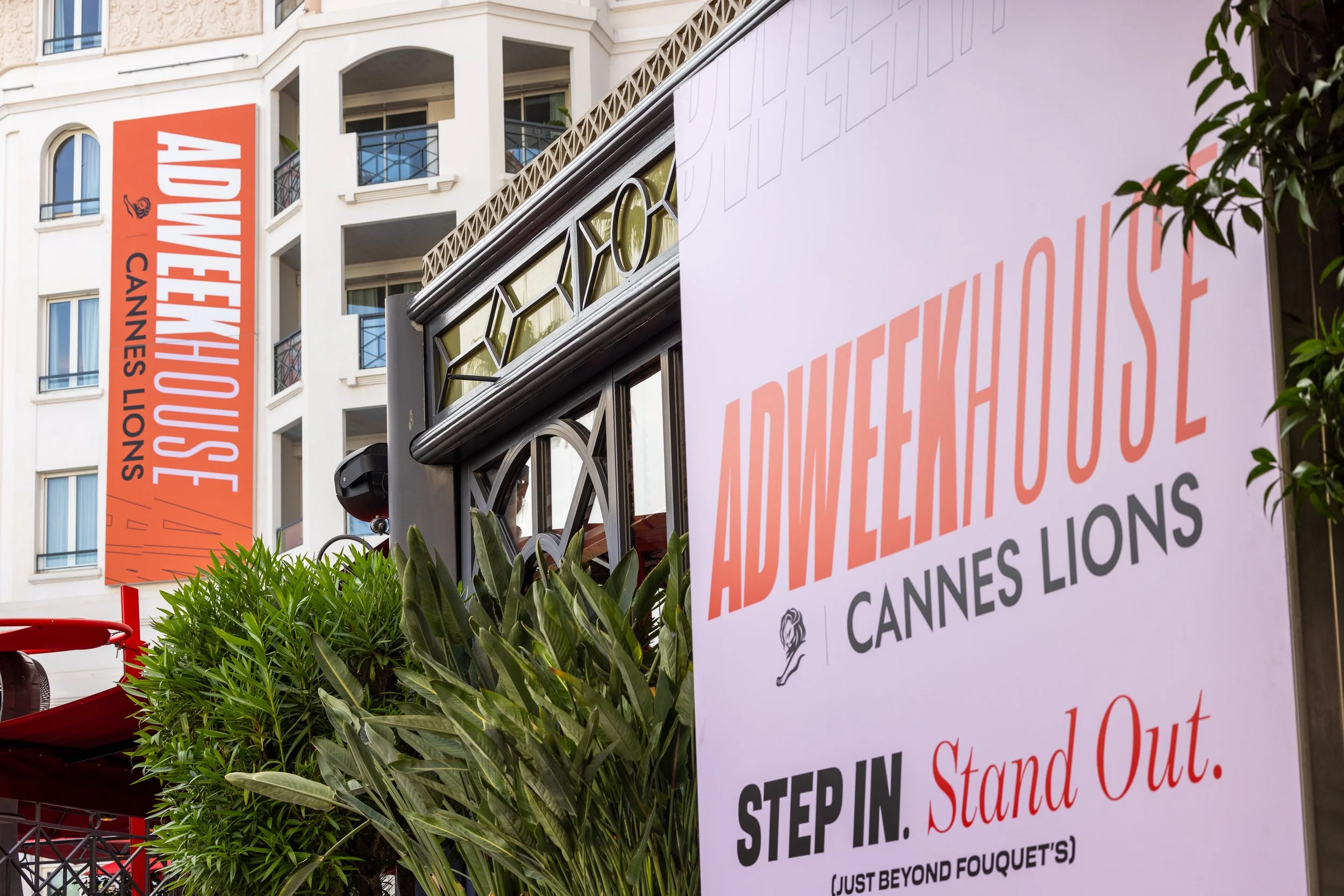 Signs for ADWEEK House at Cannes Lions event, with a building in the background and green plants in the foreground.