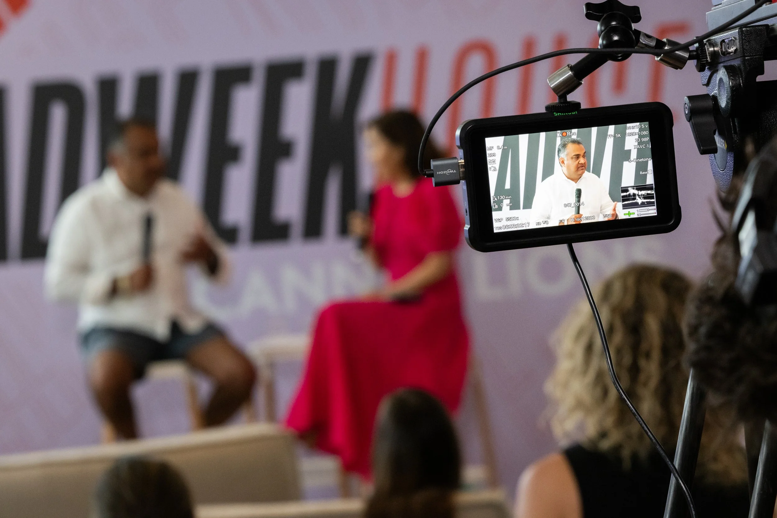 A camera captures a man and woman seated on stage during a recorded interview or discussion, with a purple backdrop and audience in the foreground.