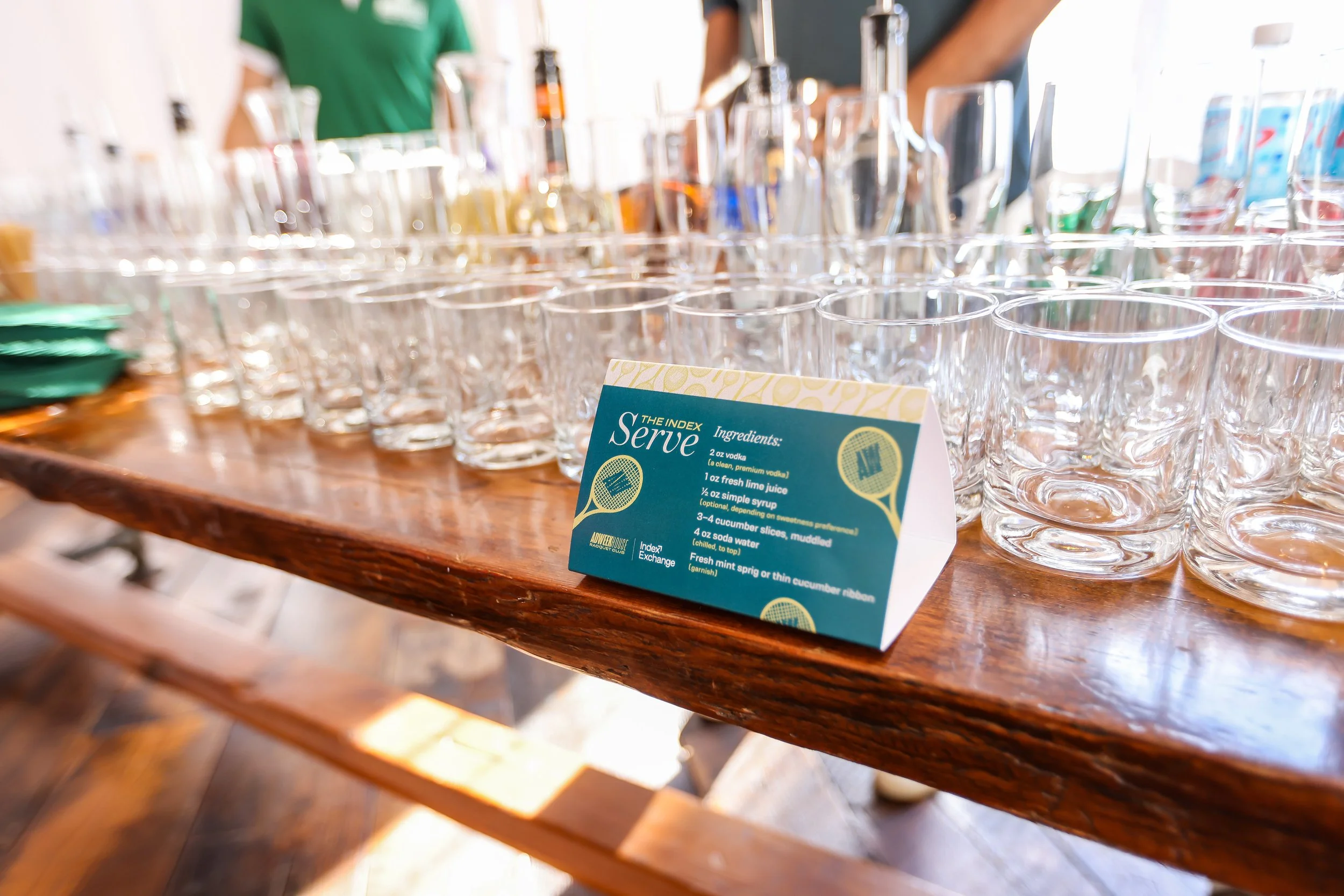 A wooden table set with multiple empty clear glasses and a small blue and yellow card with drink ingredients. The card lists ingredients for a drink called 'Serve,' including vodka, fresh lime juice, simple syrup, cucumber slices, and soda water, wit