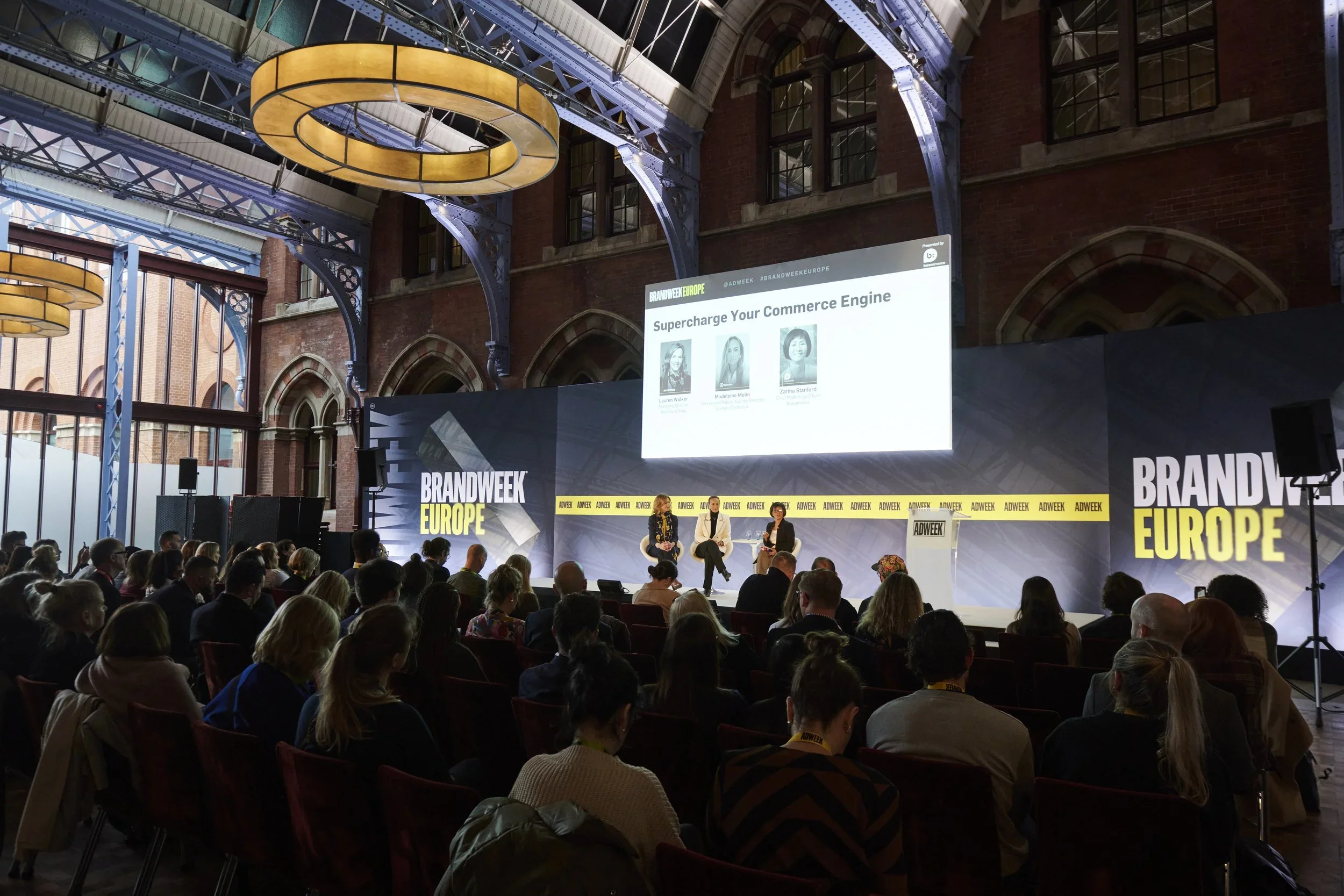An indoor conference with a stage, engaged audience, large screen displaying speakers and the text 'Supercharge Your Commerce Engine,' and banners that read 'BRANDWEEK EUROPE'.