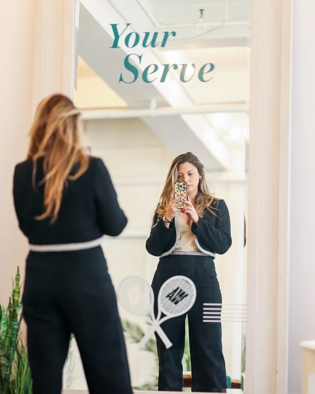 A woman taking a selfie in a mirror with the phrase "Your Serve" written above. She is dressed in a black blazer and black pants, with a beige top. The mirror features tennis racket and ball graphics.