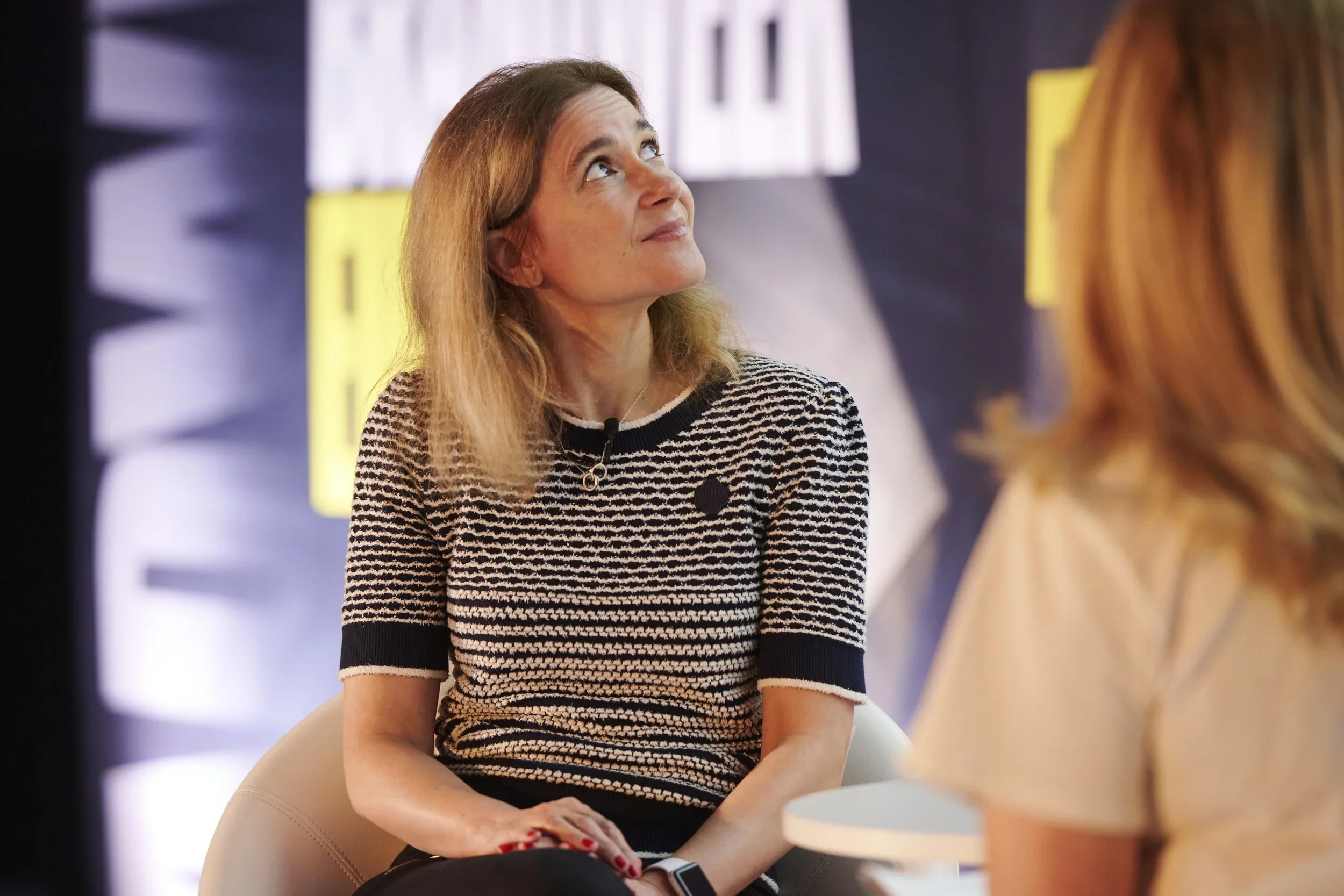 A woman with shoulder-length red hair, wearing a black and white striped shirt, sitting and looking upward during a conversation or interview.