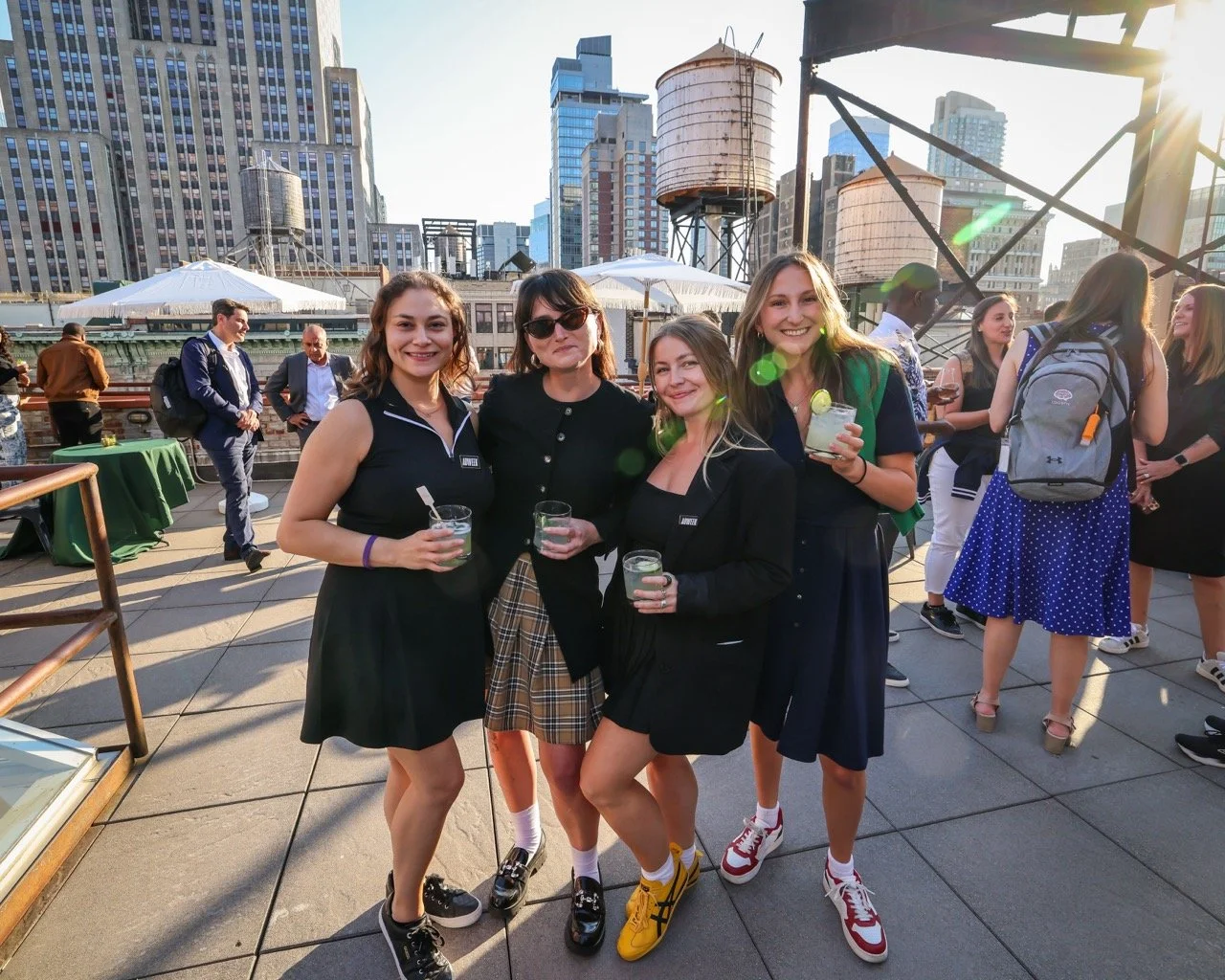 Four young women smiling and holding drinks on a rooftop with an urban cityscape in the background, during late afternoon or early evening.