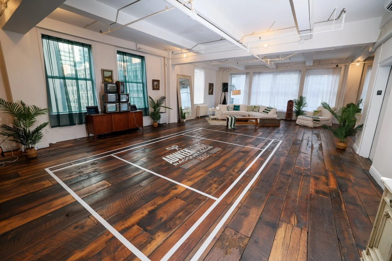 Spacious living room with hardwood floors, large windows with teal curtains, and vintage furniture. The floor has a marked rectangle with the words "ADWEEK RACQUET CLUB" There are potted plants, a TV setup, and a white sofa with green cushions.
