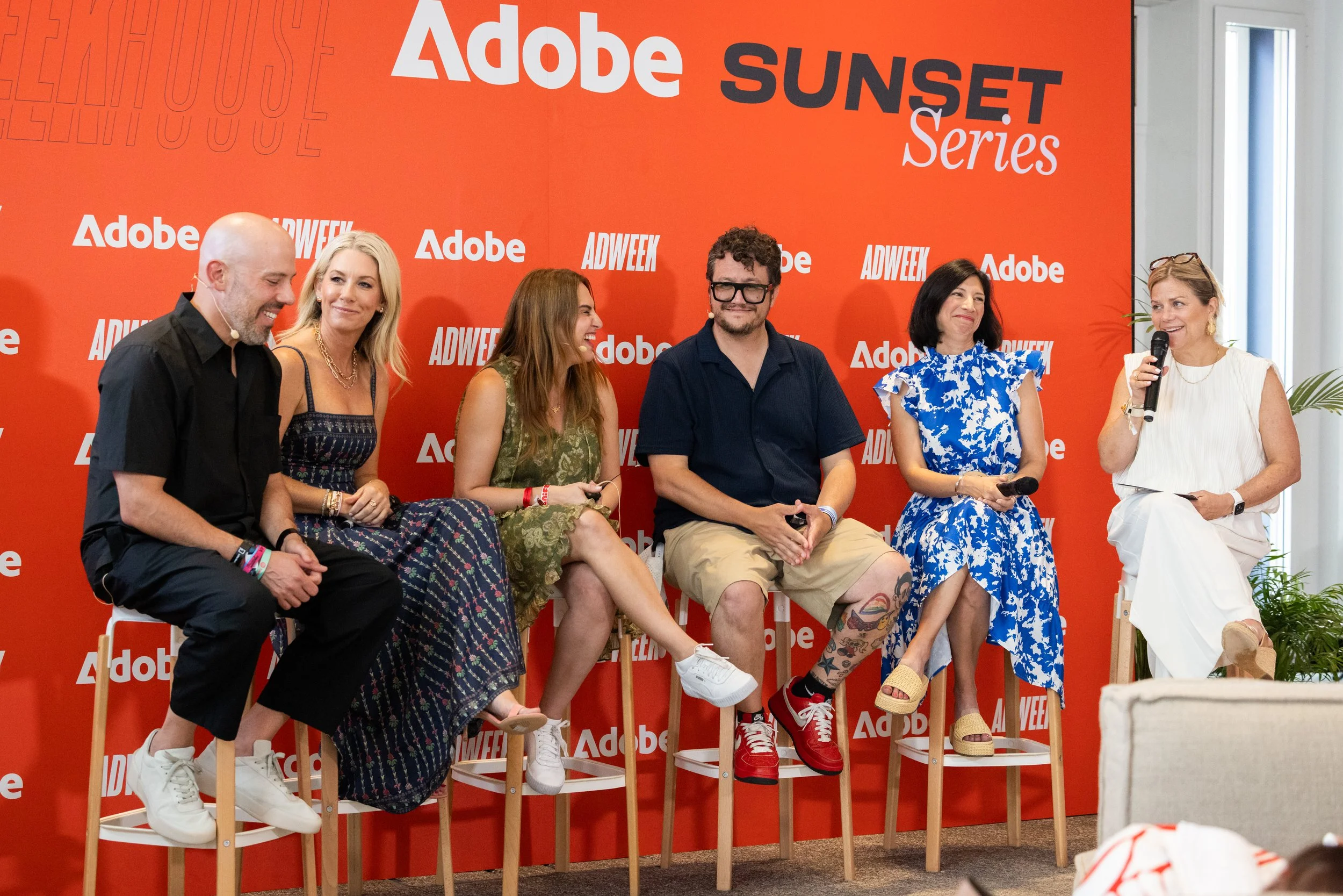 Six people sitting on chairs in front of a red backdrop with white and black text. One woman on the far right holding a microphone and speaking.