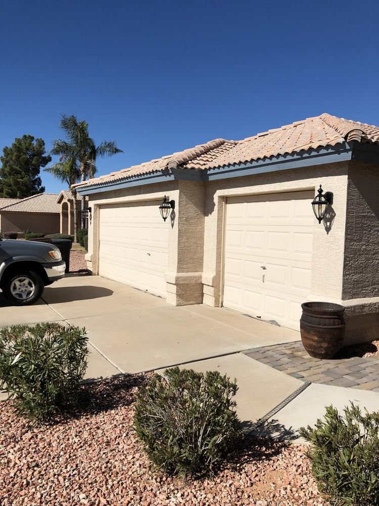 A residential house with a beige stucco exterior, tiled roof, two white garage doors, black outdoor lanterns, a black barrel planter, parked car, desert landscaping with small bushes, and a clear blue sky.