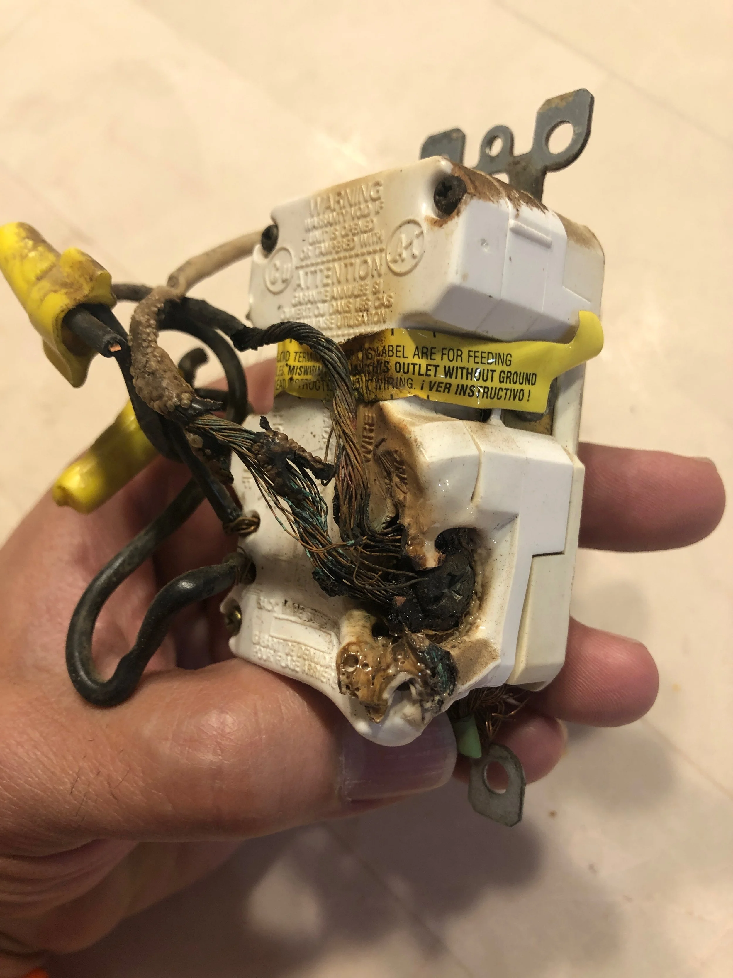 A hand holding a damaged, burnt, and charred electrical component with frayed wires and burnt plastic.