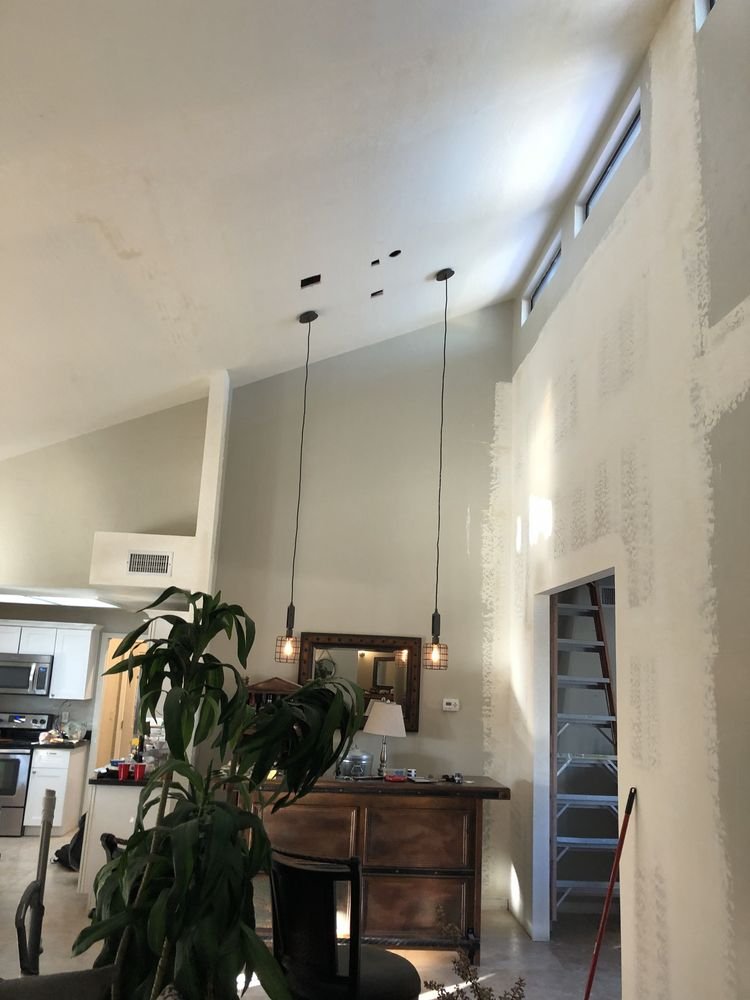 Interior view of a living space with a high ceiling, two hanging pendant lights, a wooden sideboard, a mirror, a small table lamp, and part of a kitchen visible to the left. There is a door frame on the right with a ladder inside.