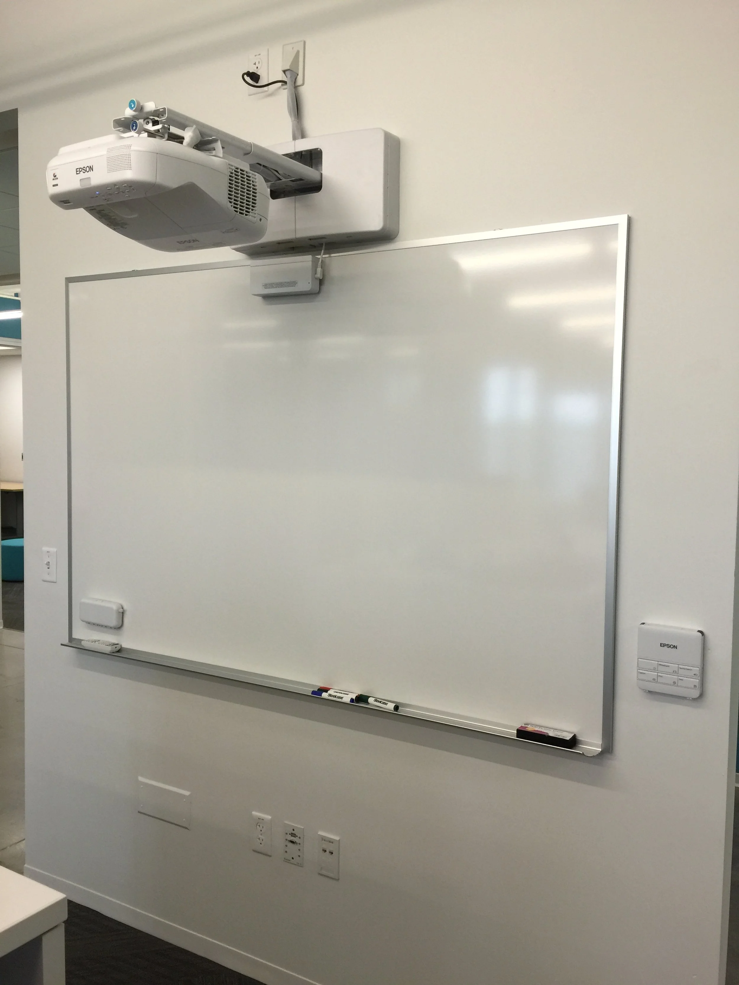 Whiteboard with markers and eraser, mounted on a white wall, with a ceiling-mounted Epson projector above, and various outlets and switches on the wall.