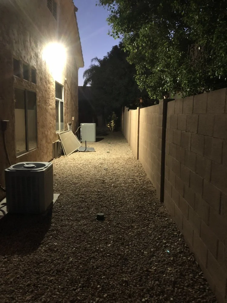 Backyard alleyway at night with gravel ground, house wall on the left, tall cinder block fence on the right, two air conditioning units, an outdoor vent, a window, and some trees with green leaves.