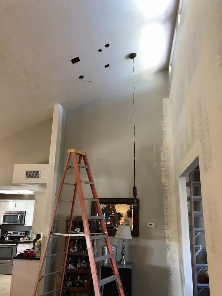 Interior view of a room under renovation with a ladder, a hanging light fixture, and a wall with unfinished paint. Kitchen visible on the left, and a mirror on the wall behind a lamp.