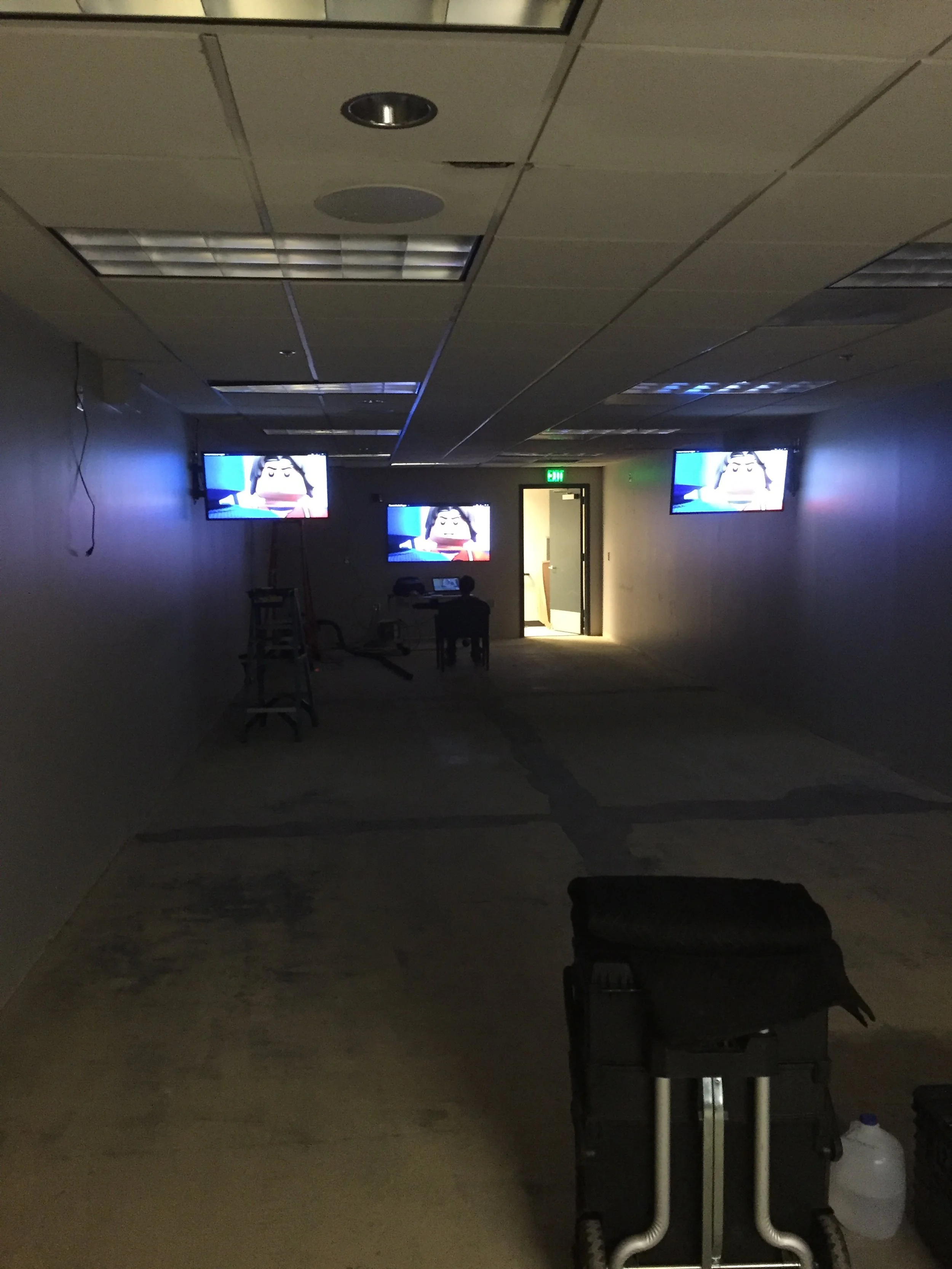 Dark room with three wall-mounted televisions showing a cartoon character, a door at the back with an illuminated green exit sign, and construction equipment and furniture in the foreground.