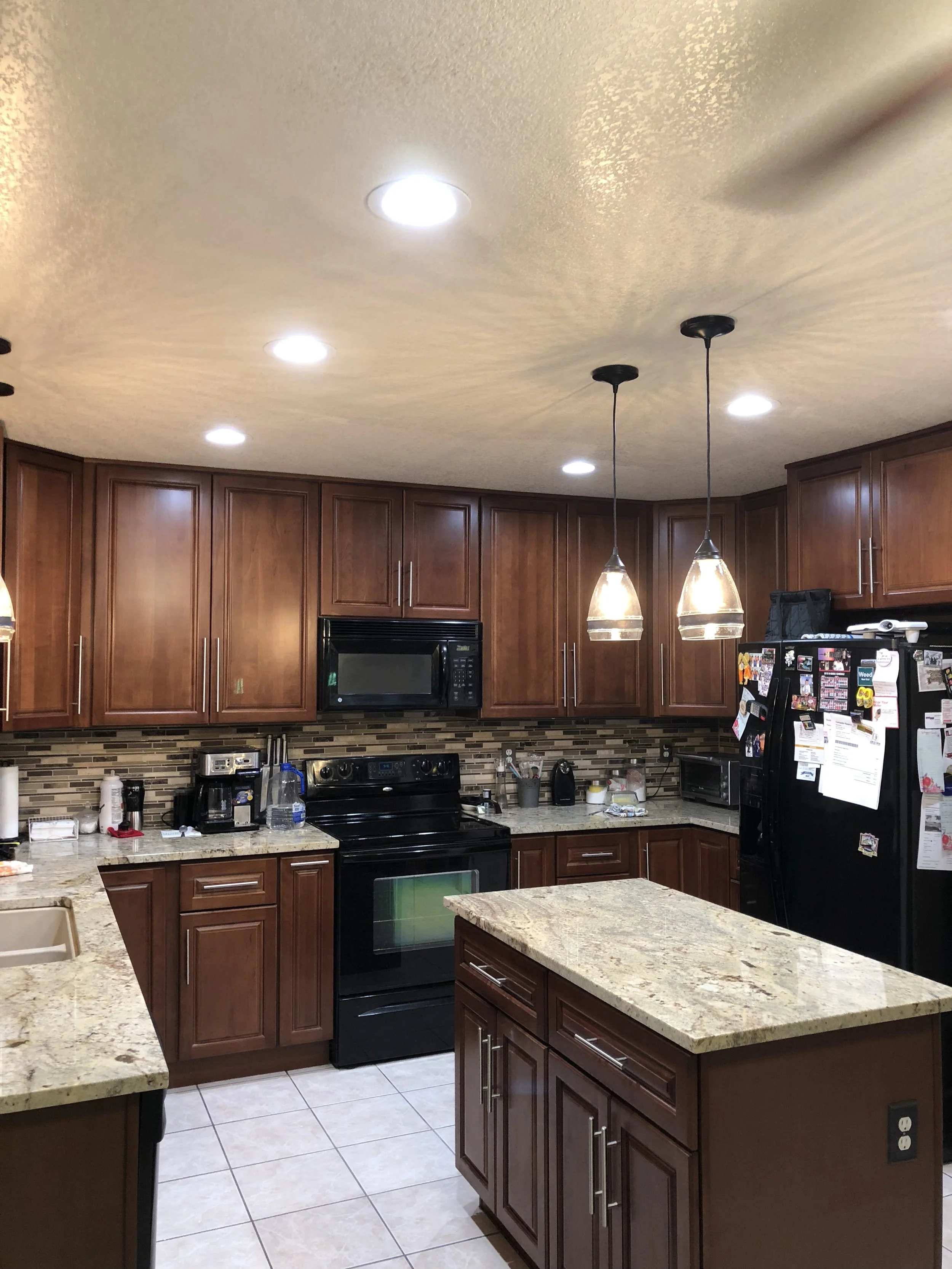 Kitchen with wooden cabinets, granite countertops, and black appliances, including a microwave, oven, and refrigerator, with two pendant lights hanging over a small island.