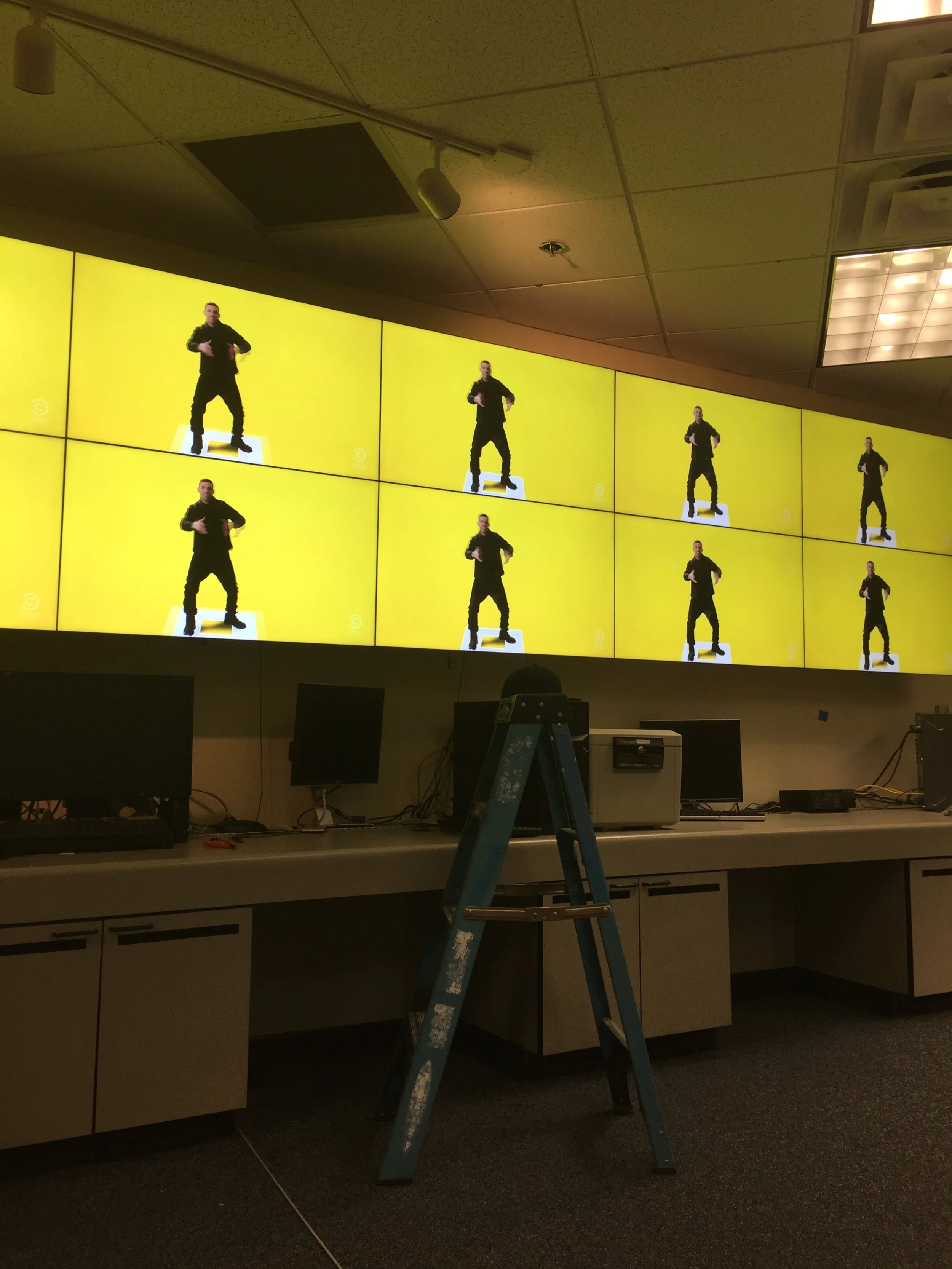 Multiple computer monitors displaying a person in black clothing dancing against a yellow background, with a ladder in the foreground and office equipment below.