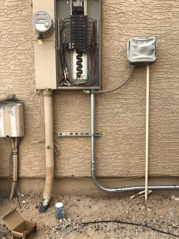 Electrical meter and breaker box on an exterior beige stucco wall, with connected conduit and wiring, and a small gray box on the right side.