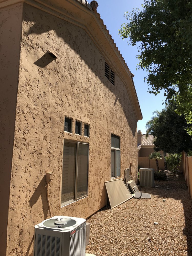 Side view of a beige stucco house with small windows, air conditioning units, and appliance parts leaning against the wall in a gravel yard, with trees and neighboring houses in the background under a sunny sky.
