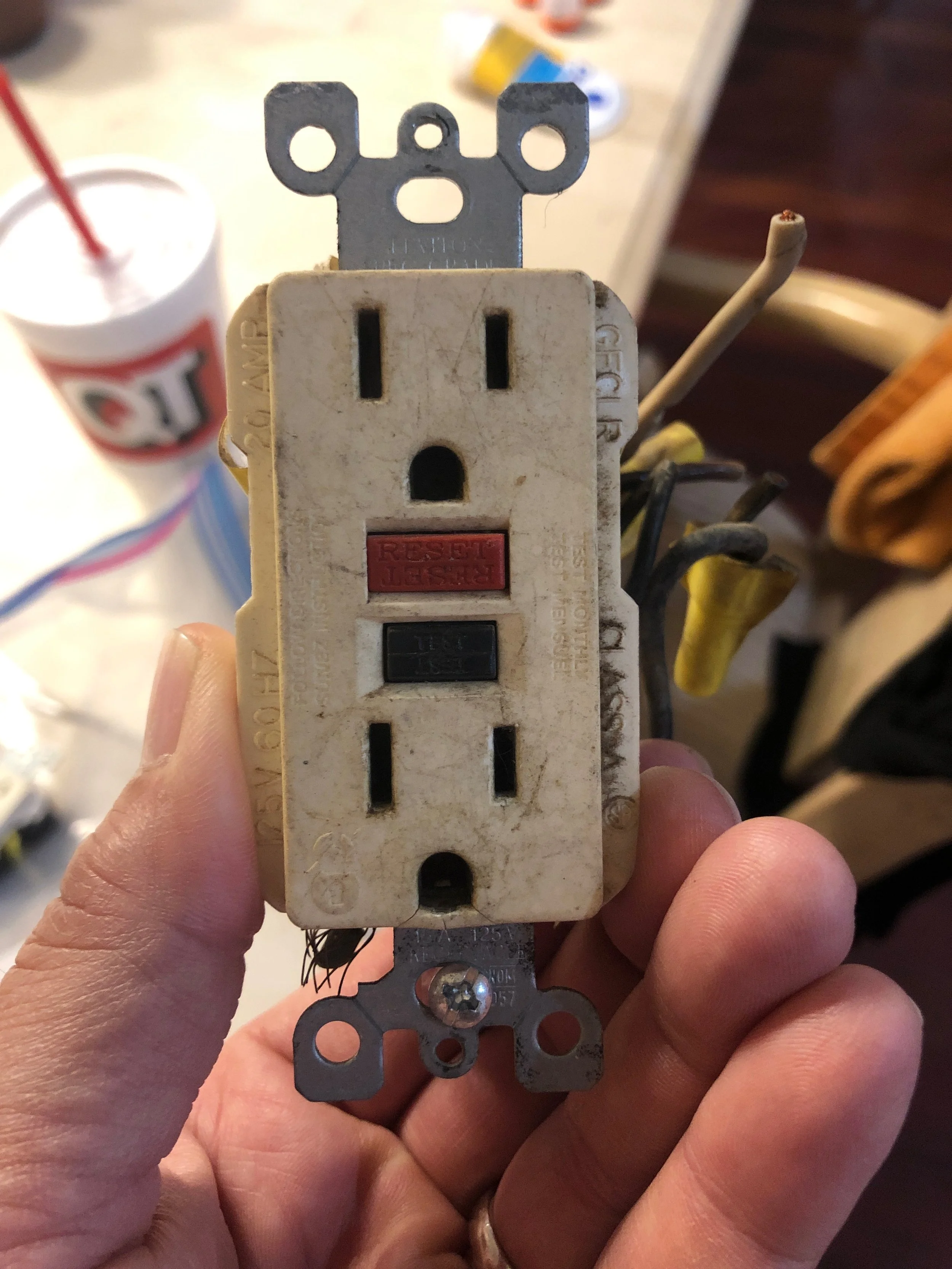 Old, worn-out electrical power outlet with a reset button, held in hand. In the background, a cup with a straw and other miscellaneous items are visible on the table.