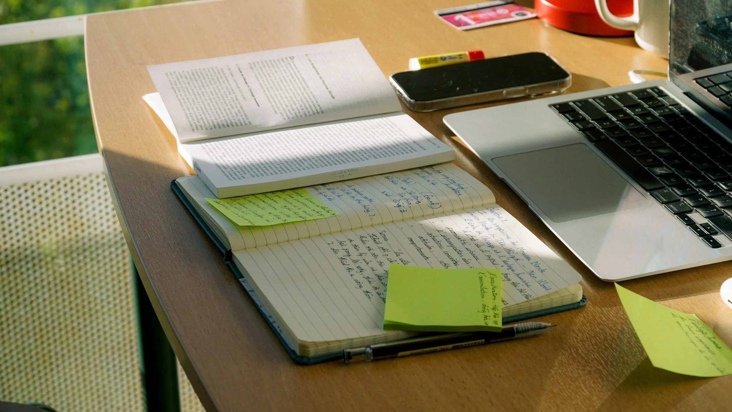 Study workspace with open notebooks, sticky notes, a pen, a smartphone, and a silver laptop on a wooden table, sunlight illuminating the scene.