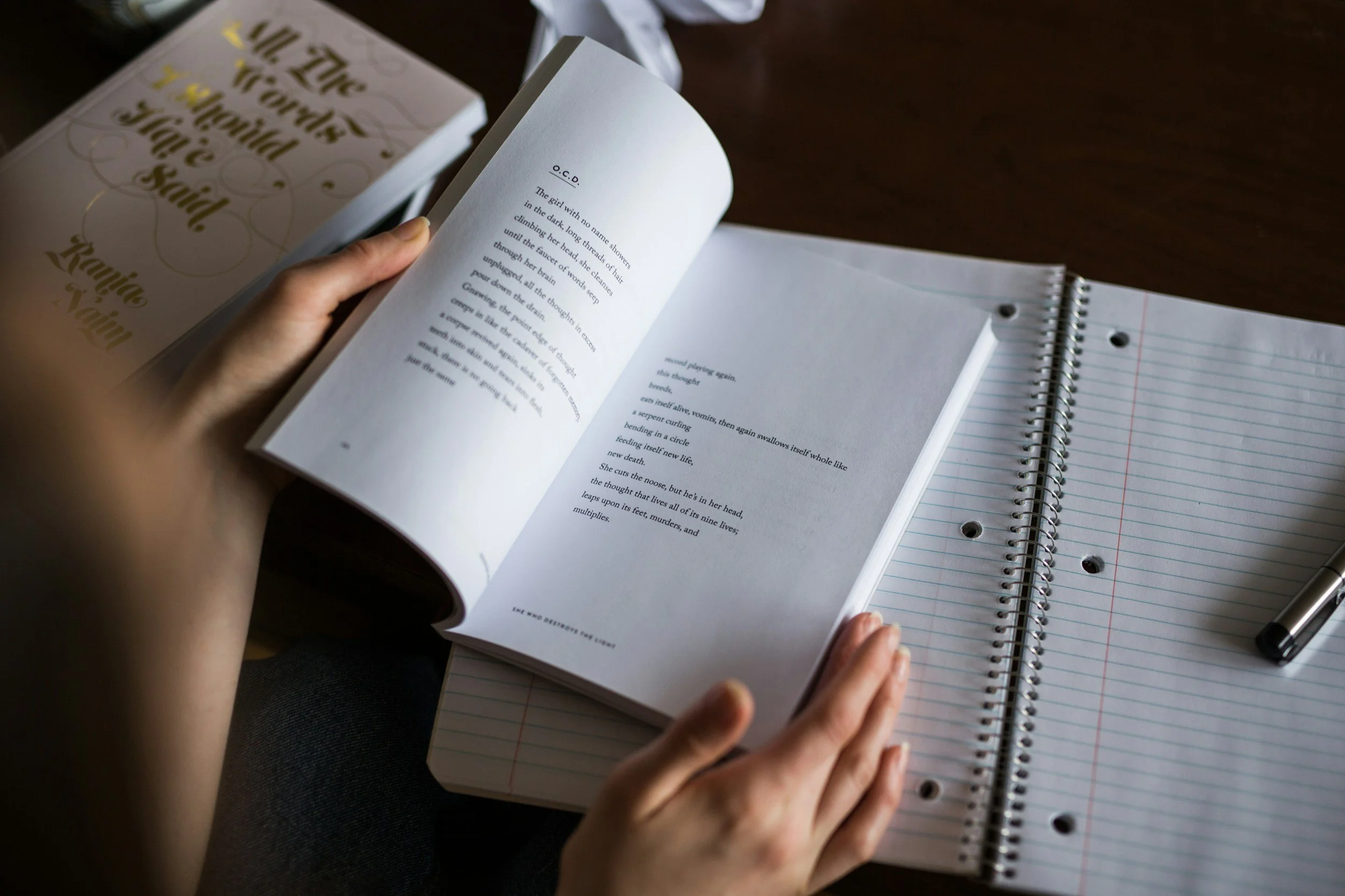 Person holding an open book with a notebook and pen on a dark wooden surface.