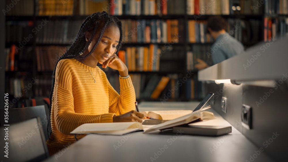 A young woman with braided hair wearing a yellow sweater, studying and writing notes at a library table with a laptop, books, and a pager in front of her. A person in the background is browsing books on a shelf.