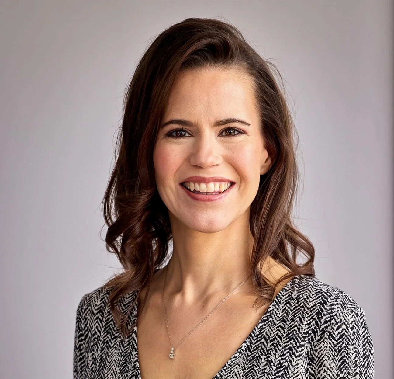 A smiling woman with wavy brown hair, wearing a black and white patterned top and a silver necklace.