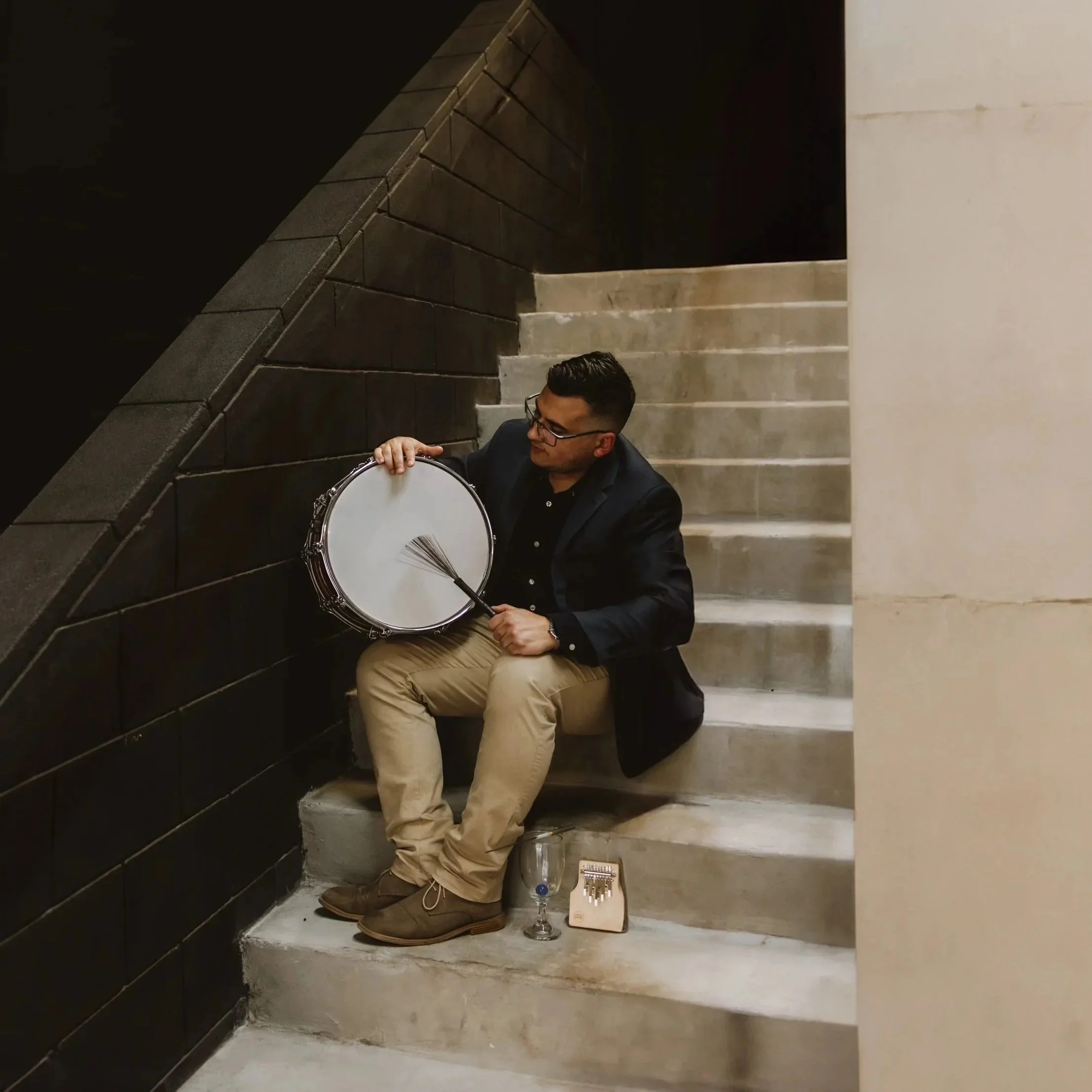A man sitting on stairs, playing a drum with a brush, with a glass and a small box at his feet.