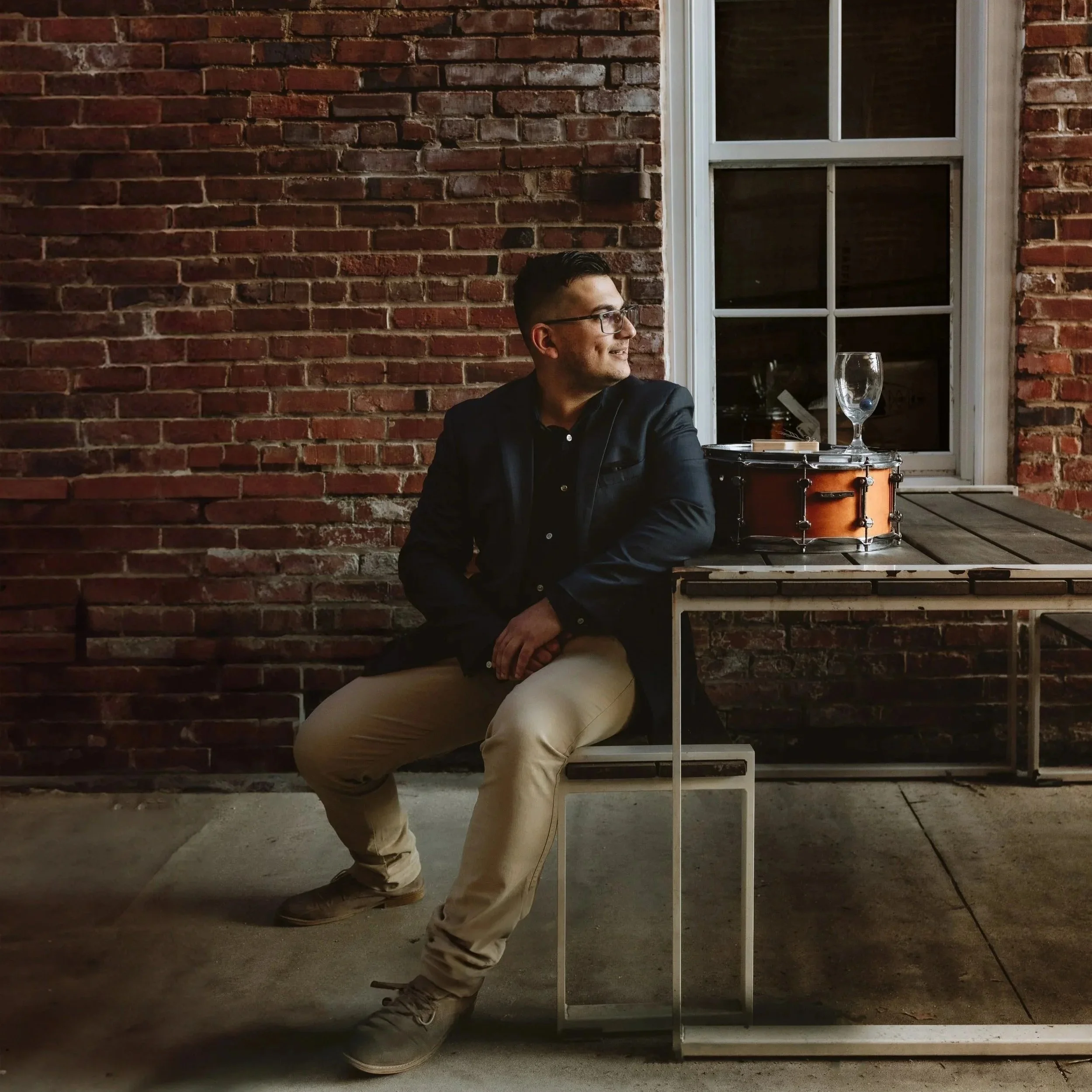 A man in glasses, wearing a dark blazer and beige pants, sitting on a bench outdoors at night, next to a table with a tambourine, an empty glass, and a book against a red brick wall.