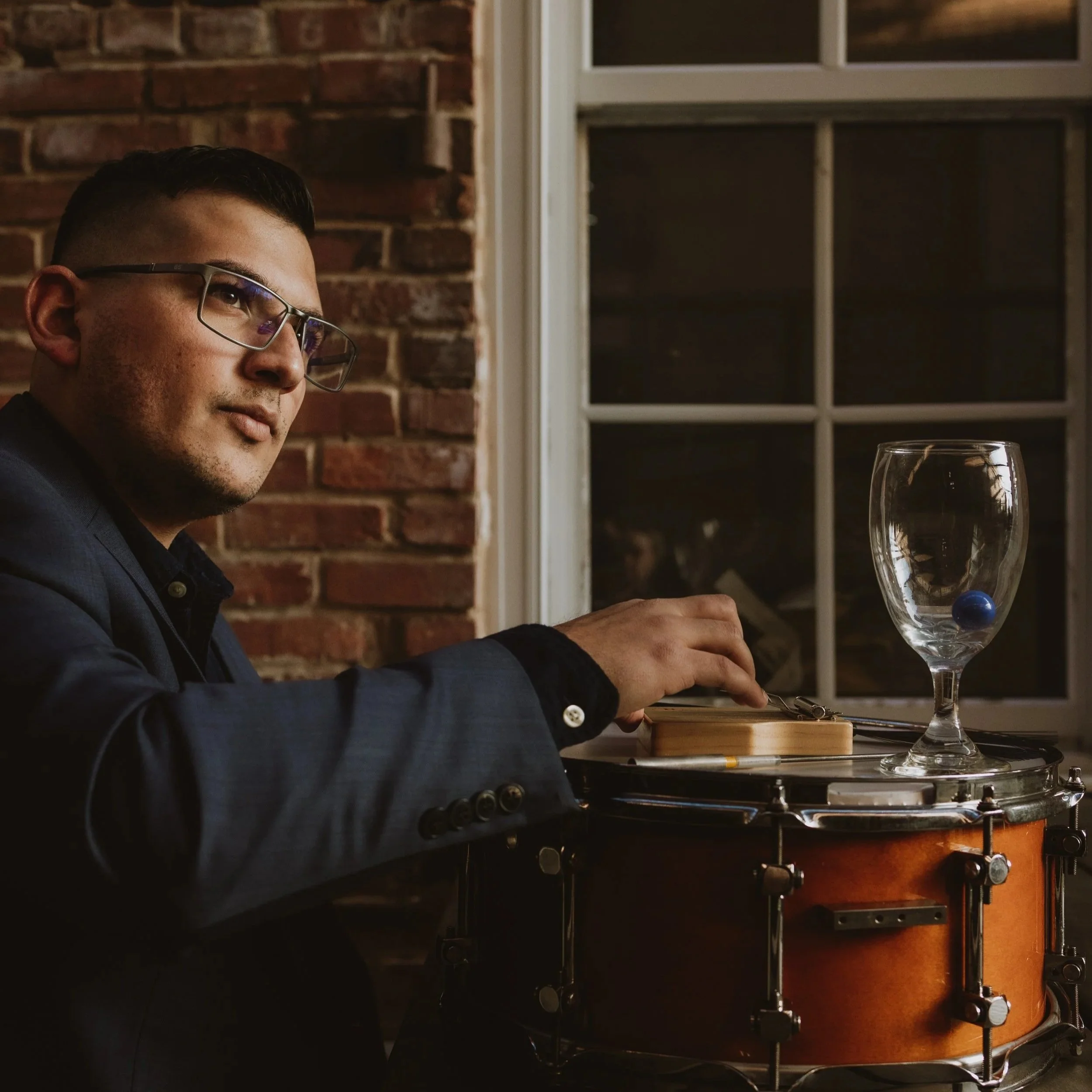 Man with glasses wearing a suit, sitting at a table with a drum, holding drumsticks with a glass of water and a blue sphere on top, in a room with brick wall and window.