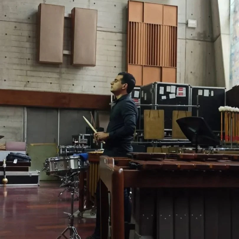 A man holding drumsticks, standing in front of a marimba in a music rehearsal room with acoustic panels on the wall.