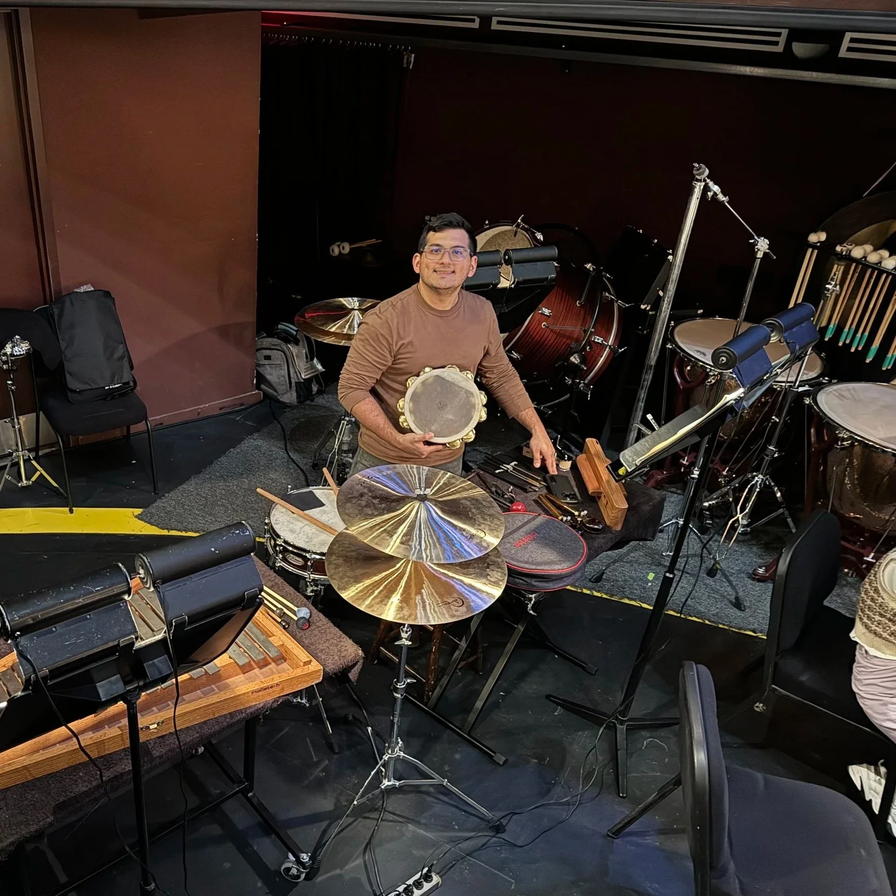 A man in a brown shirt smiling and holding a tambourine, seated at a percussion setup in a music studio surrounded by drums, cymbals, and percussion instruments.