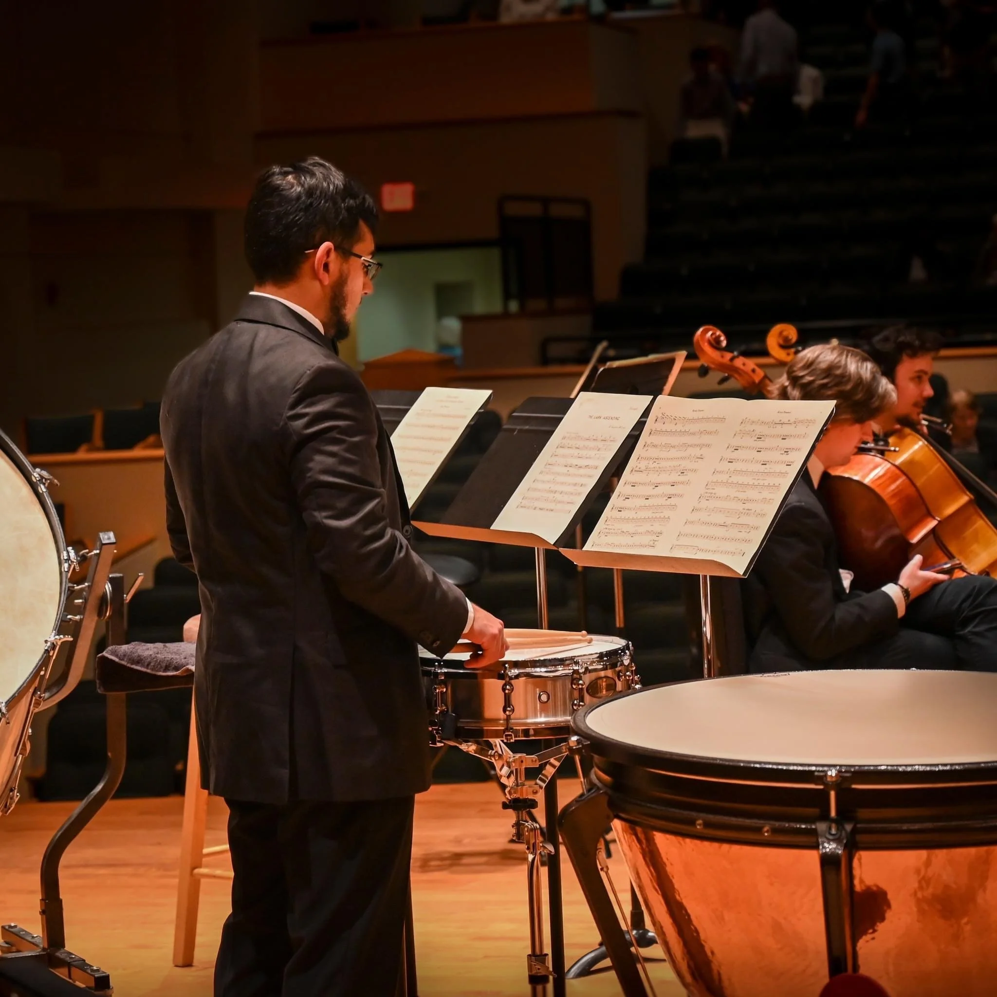 Orchestra musicians perform in a concert hall, with sheet music on stands and a percussionist at his drums, dressed in formal black attire.