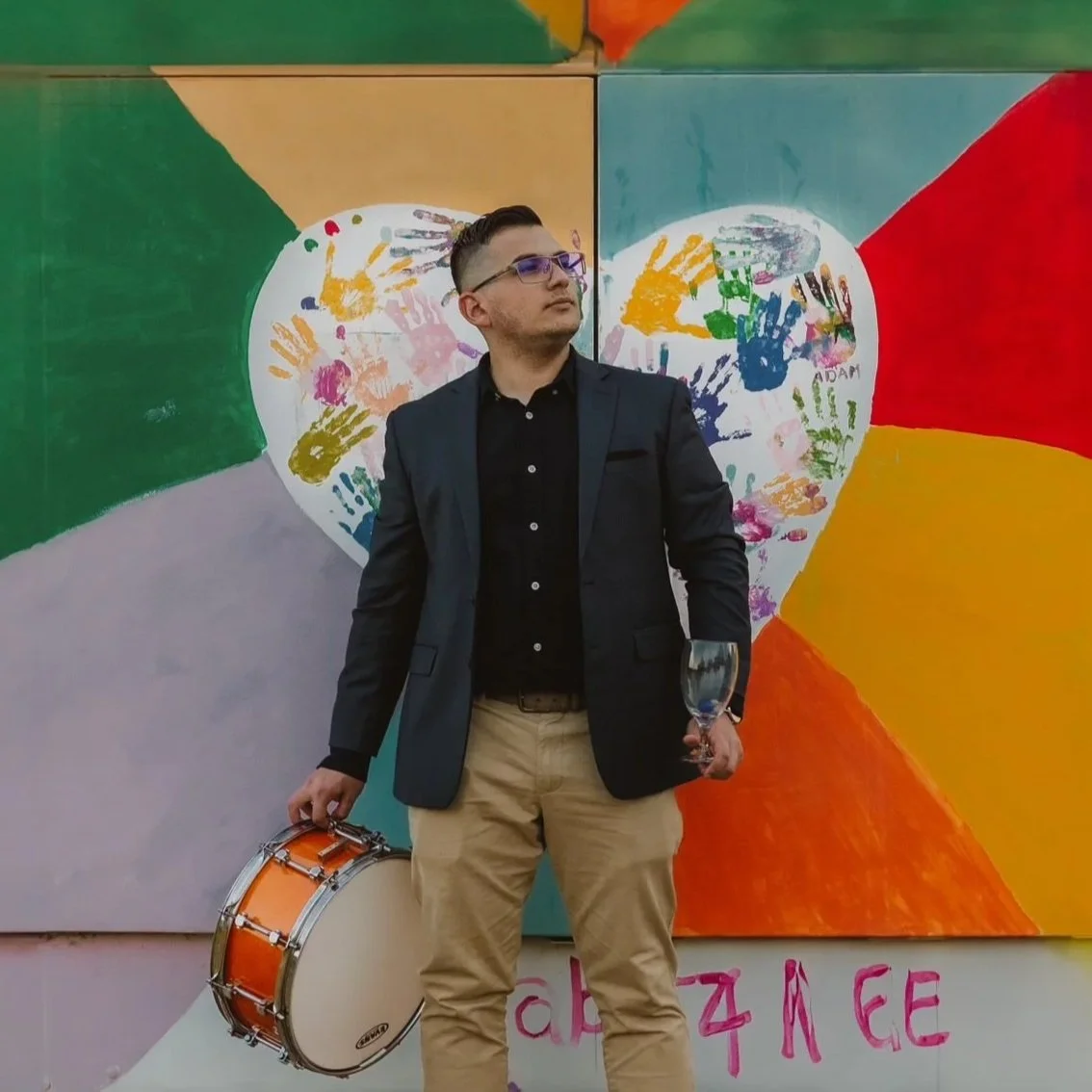 A man in a black blazer, black shirt, and khaki pants holding a glass of wine and a drum stands in front of a colorful mural with handprints and a large heart.