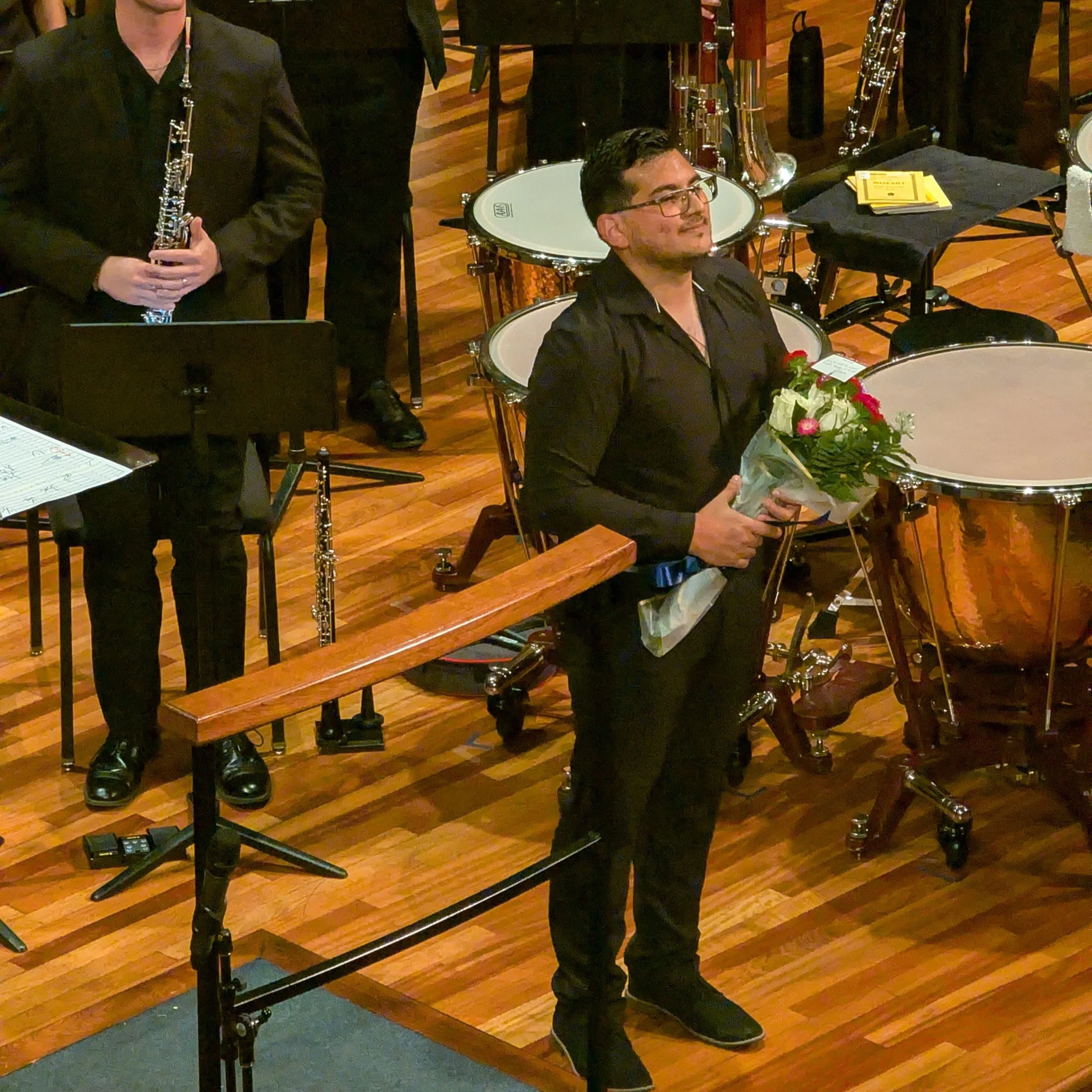 A man in a black shirt holding a bouquet of flowers on a wooden stage surrounded by percussion and wind instruments.