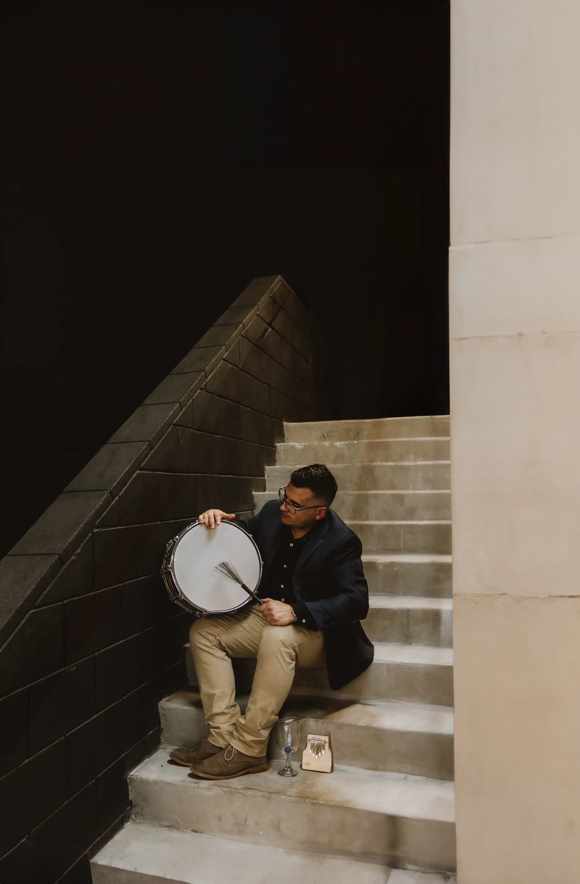 A man sitting on stairs, playing a snare drum with a drumstick in his right hand, wearing glasses, a dark blazer, black shirt, khaki pants, and brown shoes, with a tambourine, a glass with a drink, and a small musical instrument on the stairs beside him.