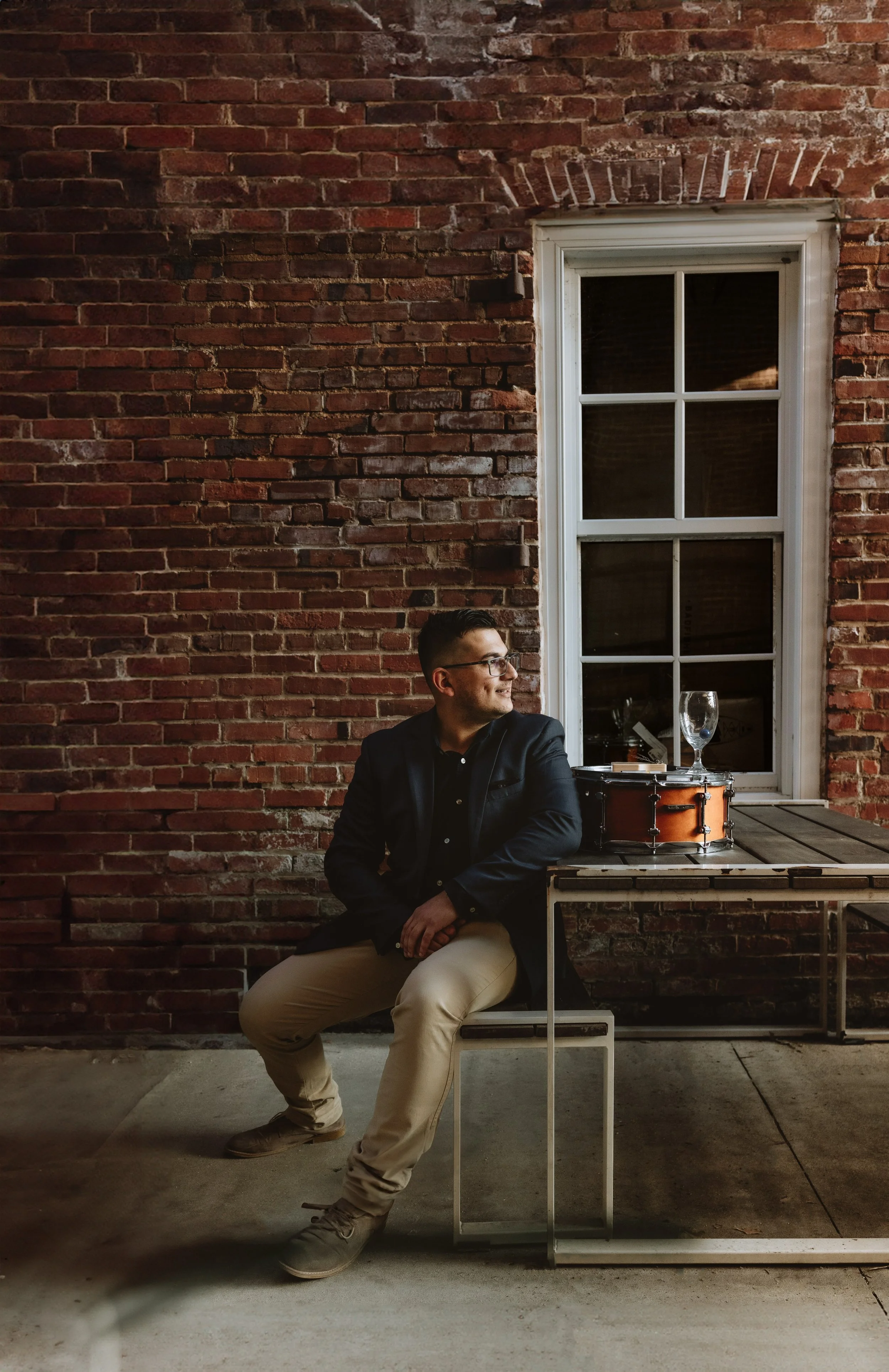 A man wearing glasses, a black jacket, and beige pants sitting on a chair outdoors, smiling, next to a table with a drum, a glass, and a small plate in front of a brick wall and window.