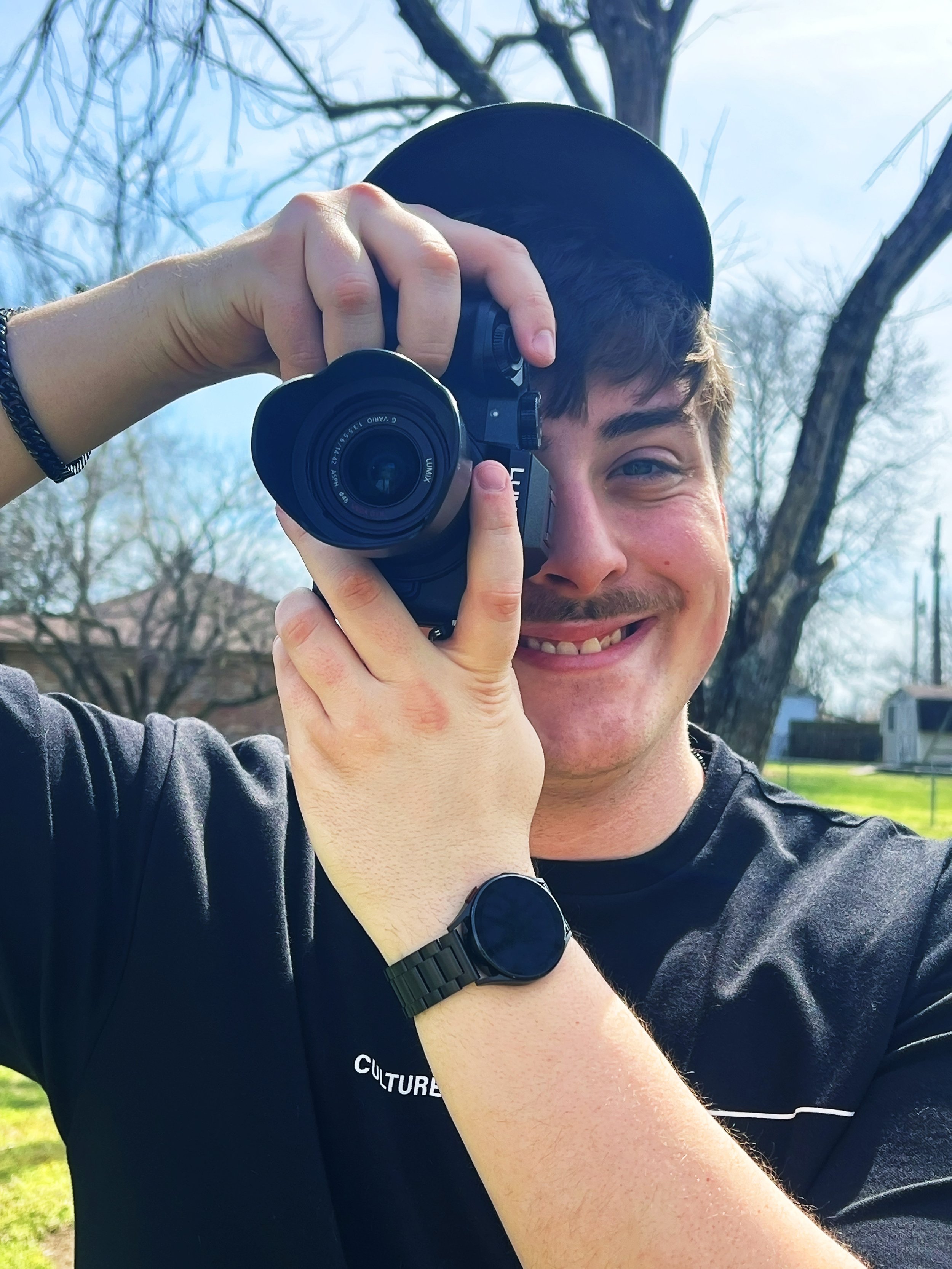 Young man smiling while taking a photo outdoors with a camera, wearing a cap, black shirt, and a smartwatch, with trees and a building in the background.