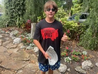 A boy standing outdoors on a rocky pathway, wearing sunglasses, a black T-shirt with a red pig graphic and text, and blue shorts, holding a crumpled newspaper, giving a thumbs-up gesture.