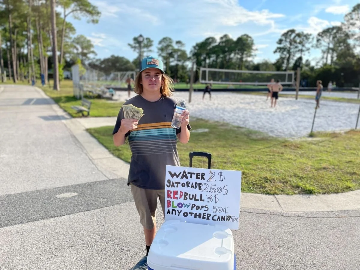 A young man standing outdoors by a beach volleyball court, holding money and a water bottle, with a cooler and a whiteboard sign listing items for sale, including water, Gatorade, Red Bull, blow pops, and other candies.