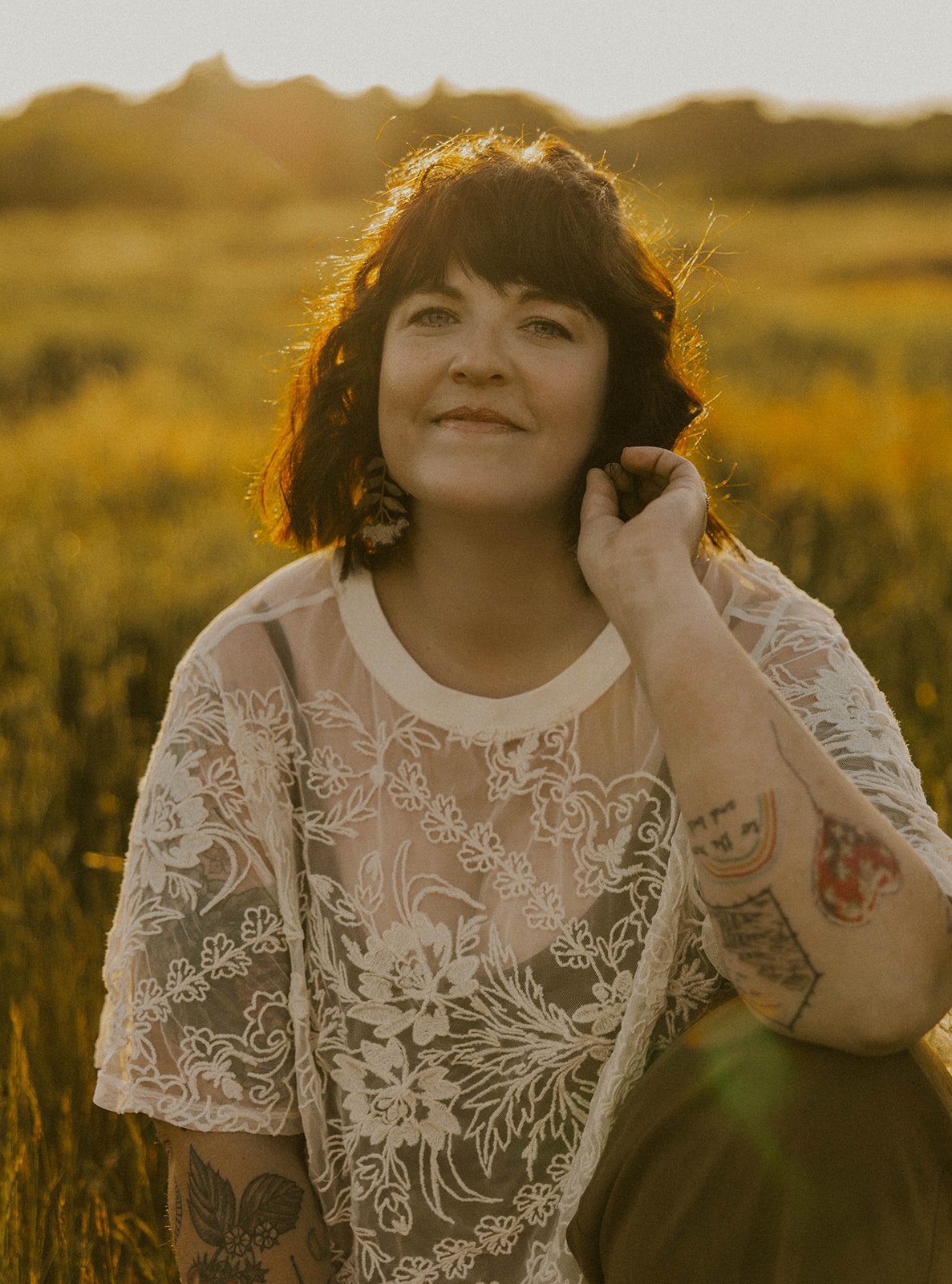 A woman with shoulder-length dark hair, wearing a white lace top, sitting outdoors in a field at sunset. She has tattoos on her arm and is smiling softly.