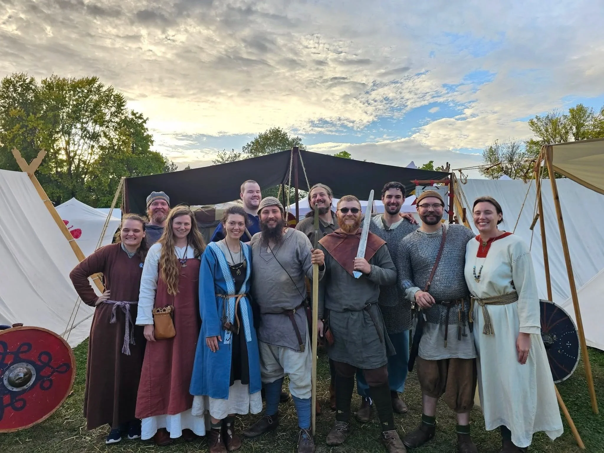 Group of people dressed in medieval costumes at a historical reenactment event outdoors, with tents and trees in the background.