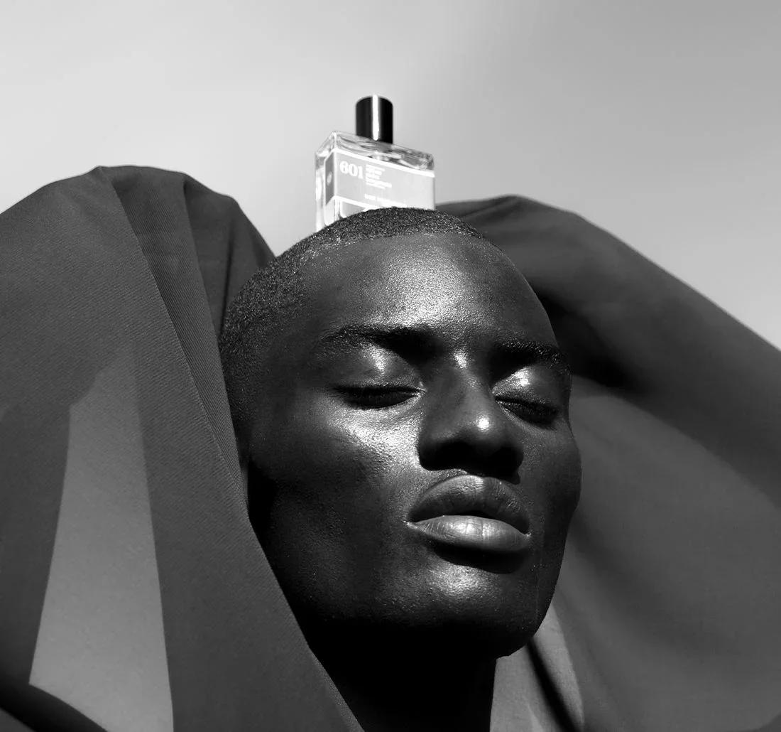 Black person with closed eyes and relaxed expression, lying down with head resting on a fabric surface, with a bottle of perfume placed on the head.