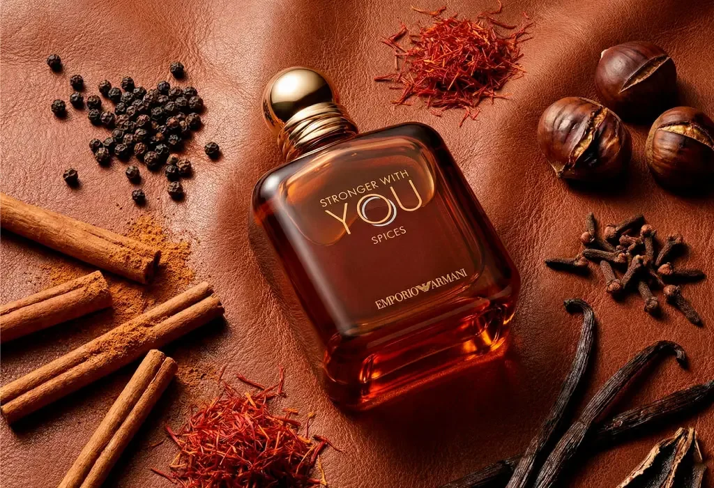 A bottle of Emporio Armani Stronger With You Spice perfume surrounded by spices, including cinnamon sticks, black peppercorns, cloves, dried saffron, vanilla beans, and chestnuts, on a brown textured background.