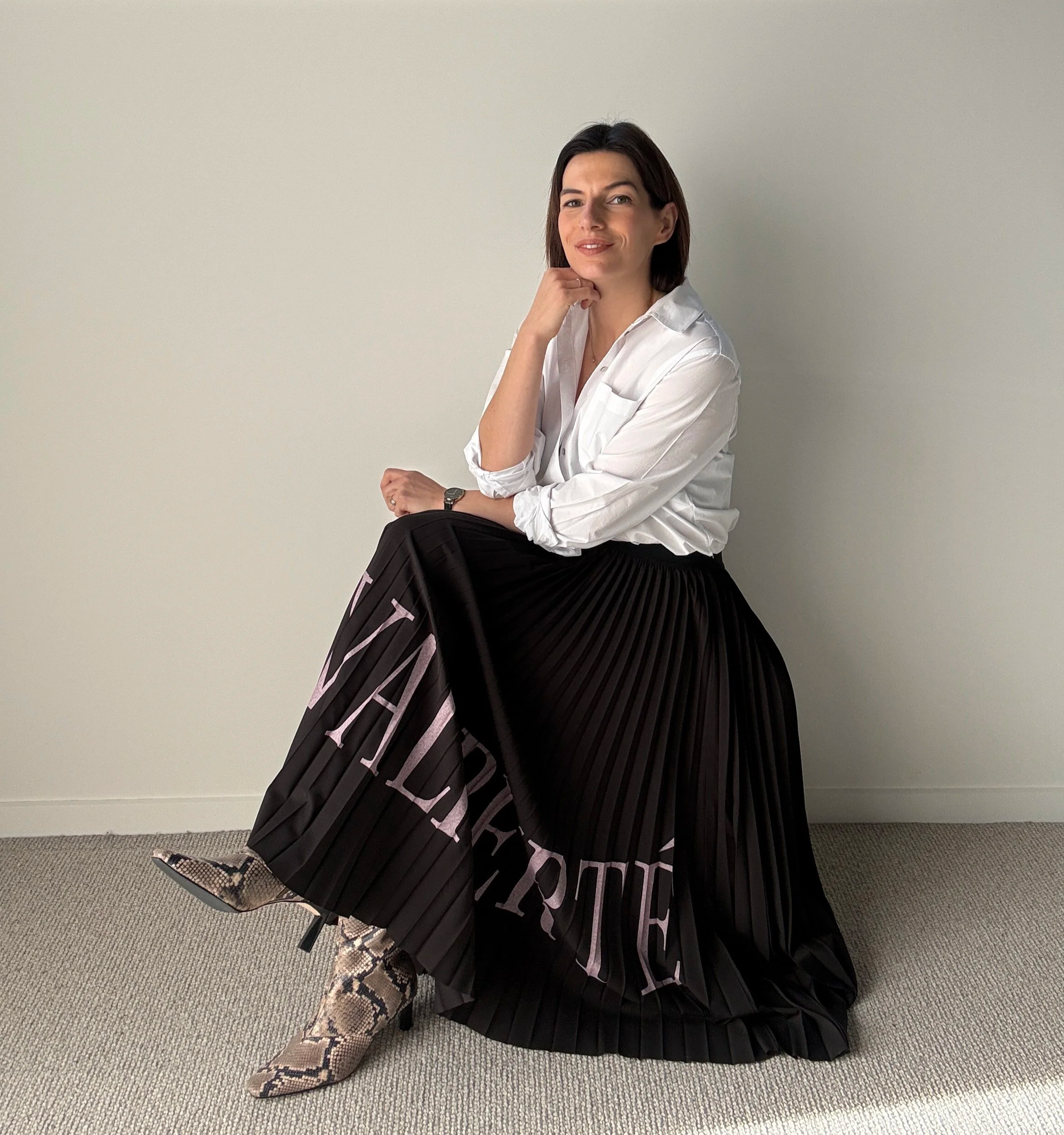 Courtney Gaudion Queenstown personal stylist sitting on the floor wearing a white blouse, black pleated skirt, snake print shoes, posing with her right hand under her chin against a plain light gray background.