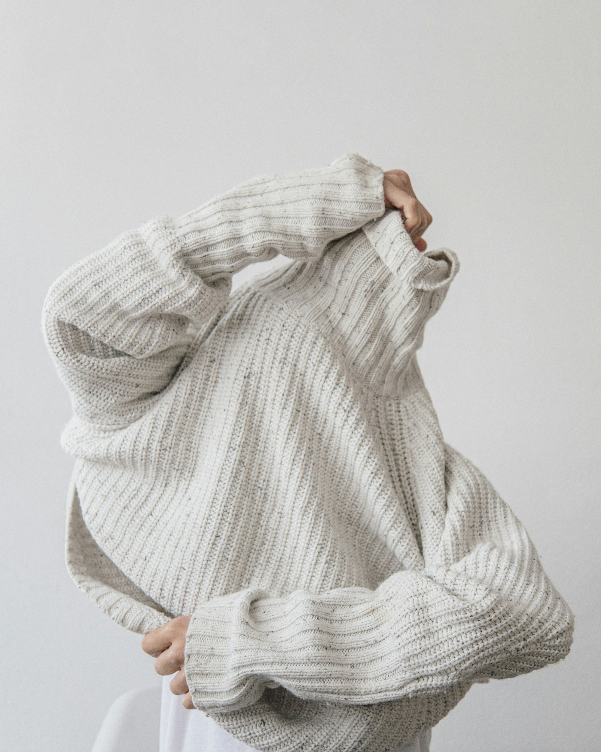 Person wearing cream-colored, chunky knit sweater with long sleeves, standing against a plain white wall.
