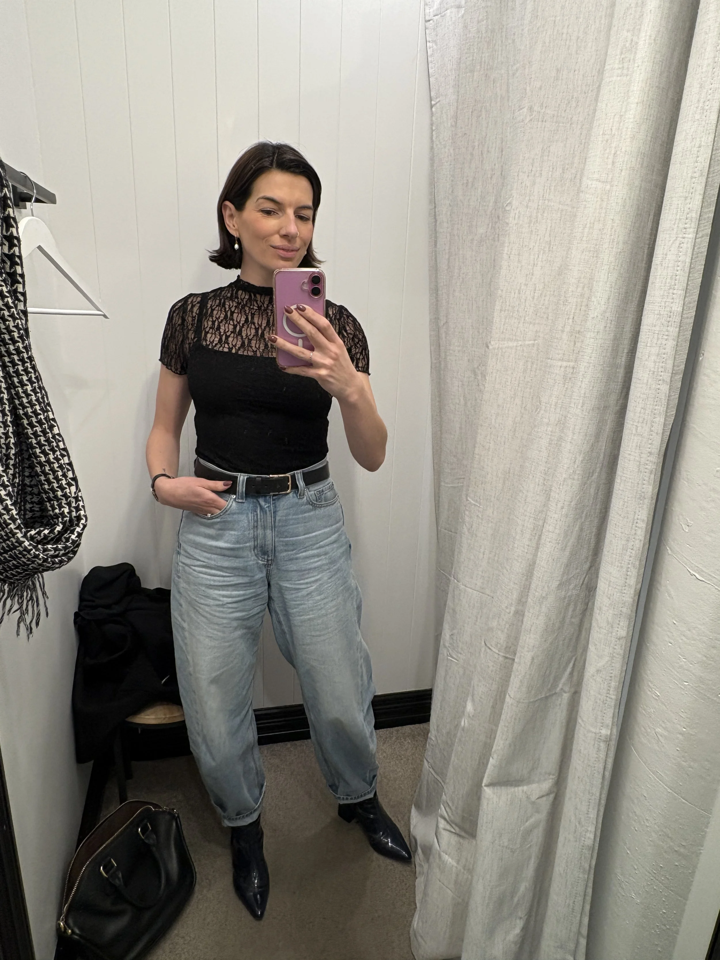 Courtney Gaudion a personal stylist wearing a black lacy top, light blue jeans, and black boots taking a mirror selfie in a fitting room with beige curtains, a black handbag, and a black jacket on the floor.