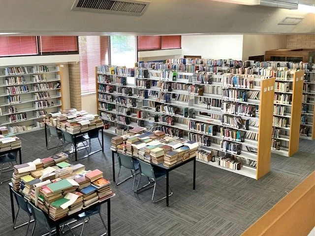 A library room filled with bookshelves and tables stacked with books.