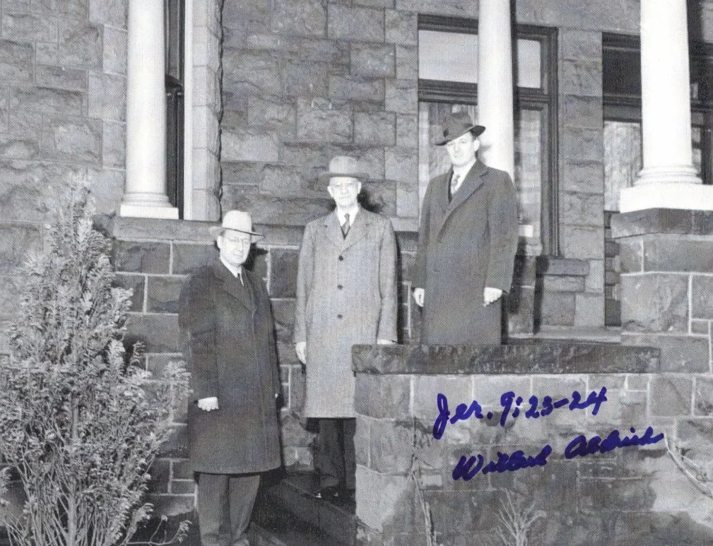 Black and white photo of three men standing outside a brick house with columns, dressed in vintage clothing and hats, with a handwritten note 'Jeremiah. 9, 28-24' and a signature.
